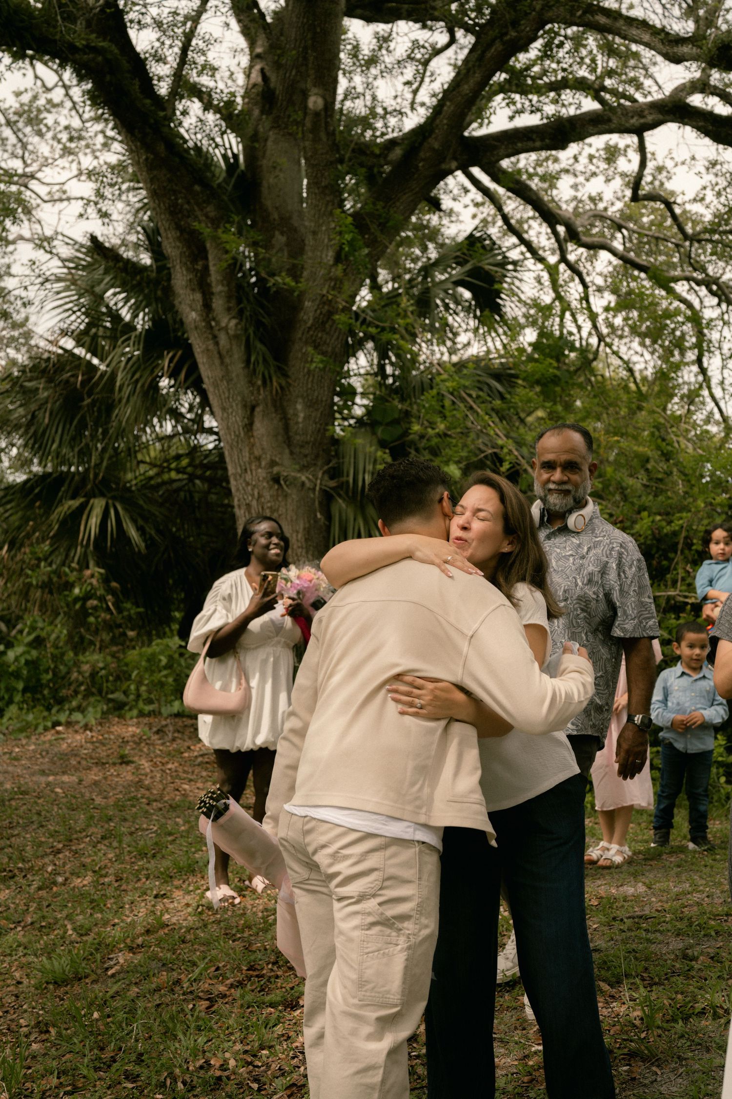 Groom in beige suit hugging a guest at an outdoor wedding ceremony under a large tree.