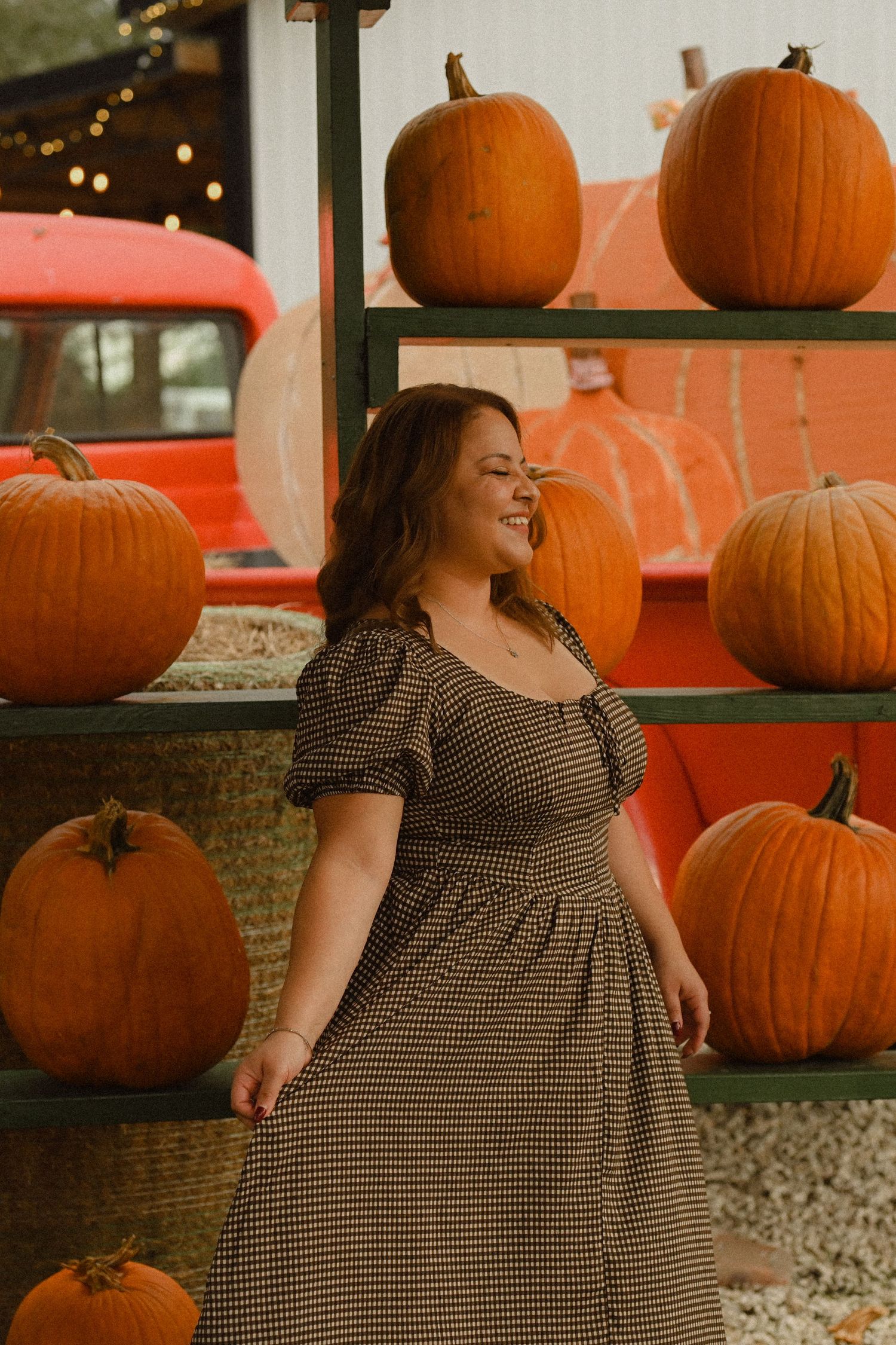 A person in a checkered dress poses at a rustic pumpkin patch display with orange pumpkins and warm autumn lighting.