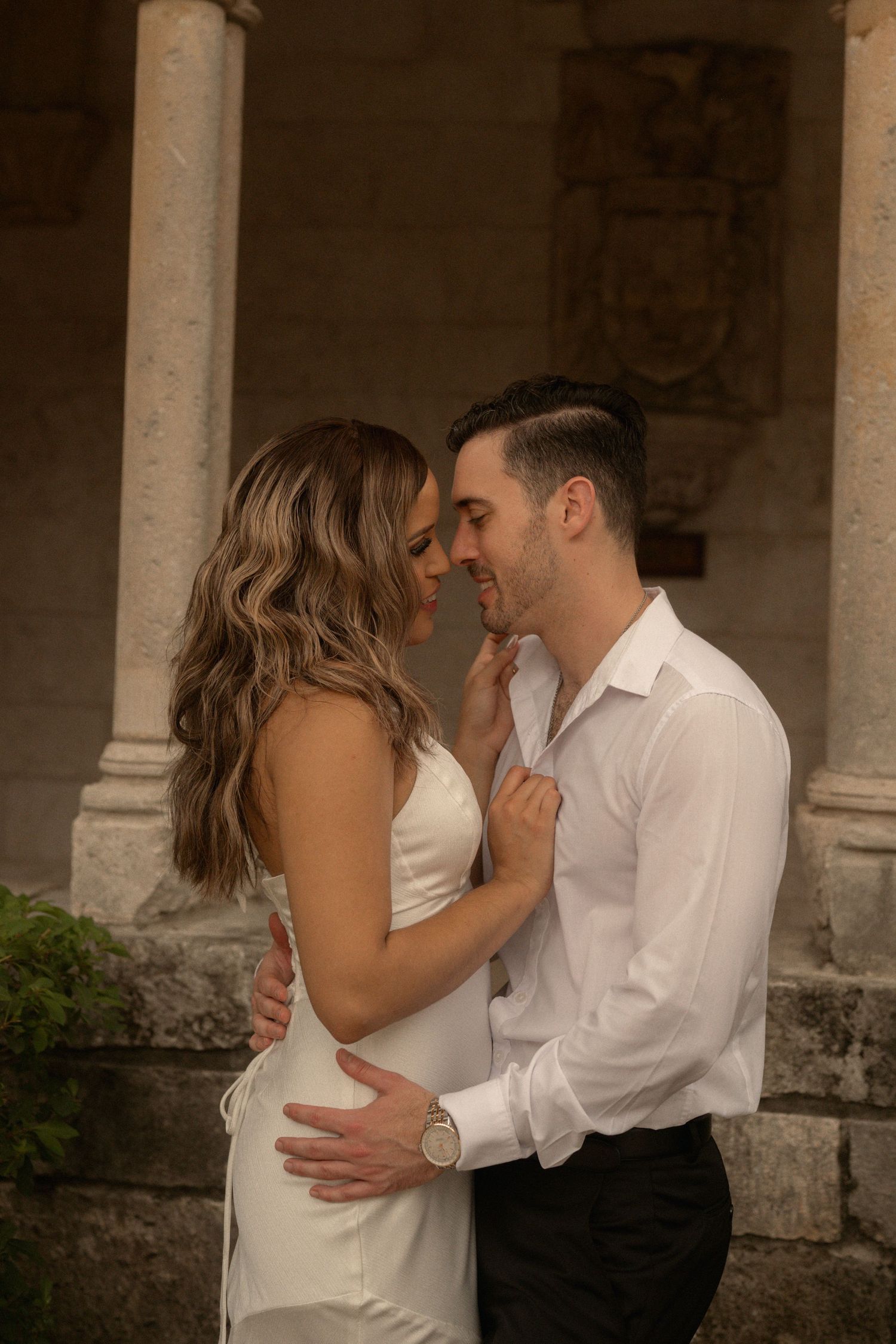 Engaged couple embracing nose to nose beside stone columns in an old-world courtyard during their photo shoot.