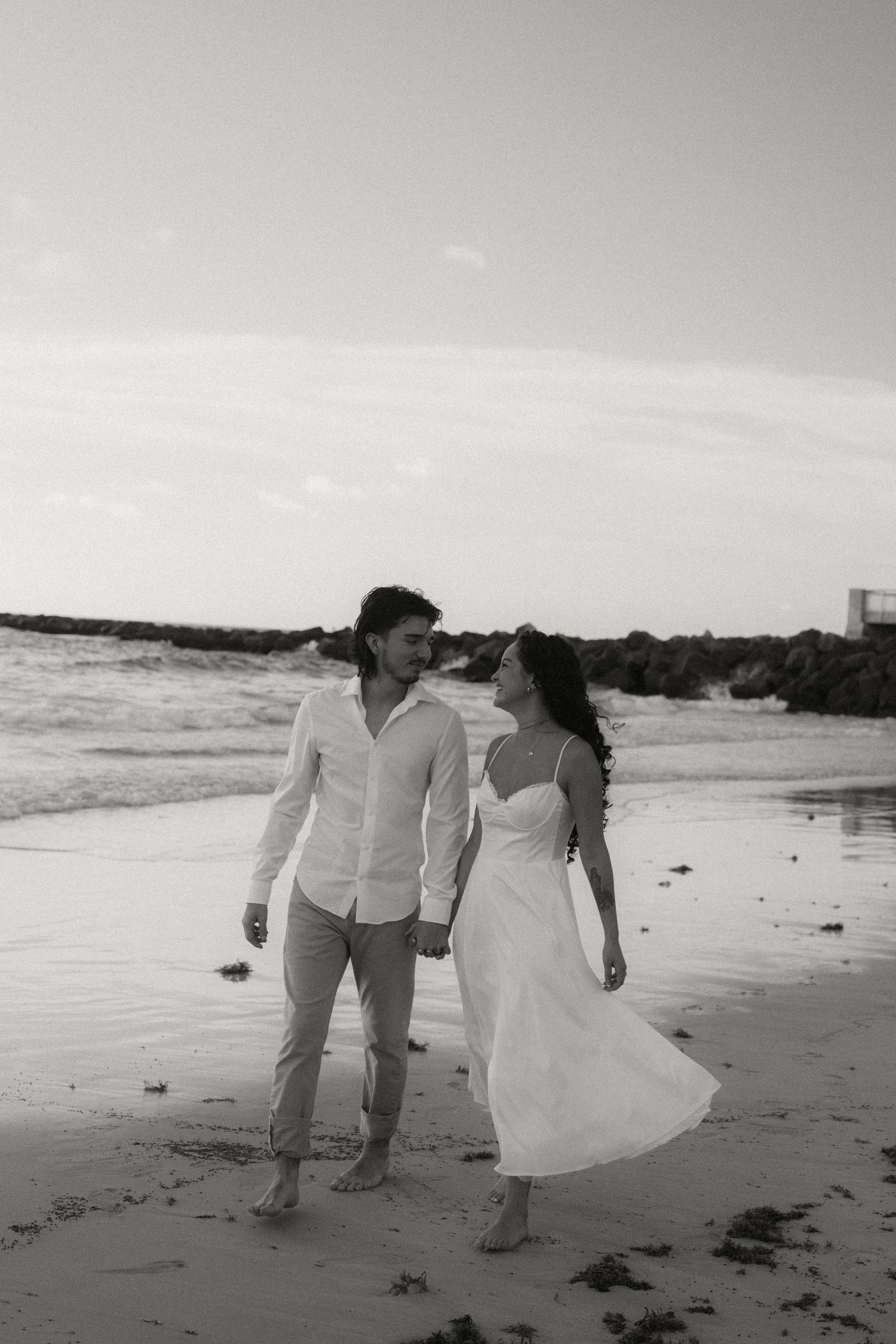 Couple walking hand in hand on beach at sunset in black and white, wearing white outfits, smiling at each other.