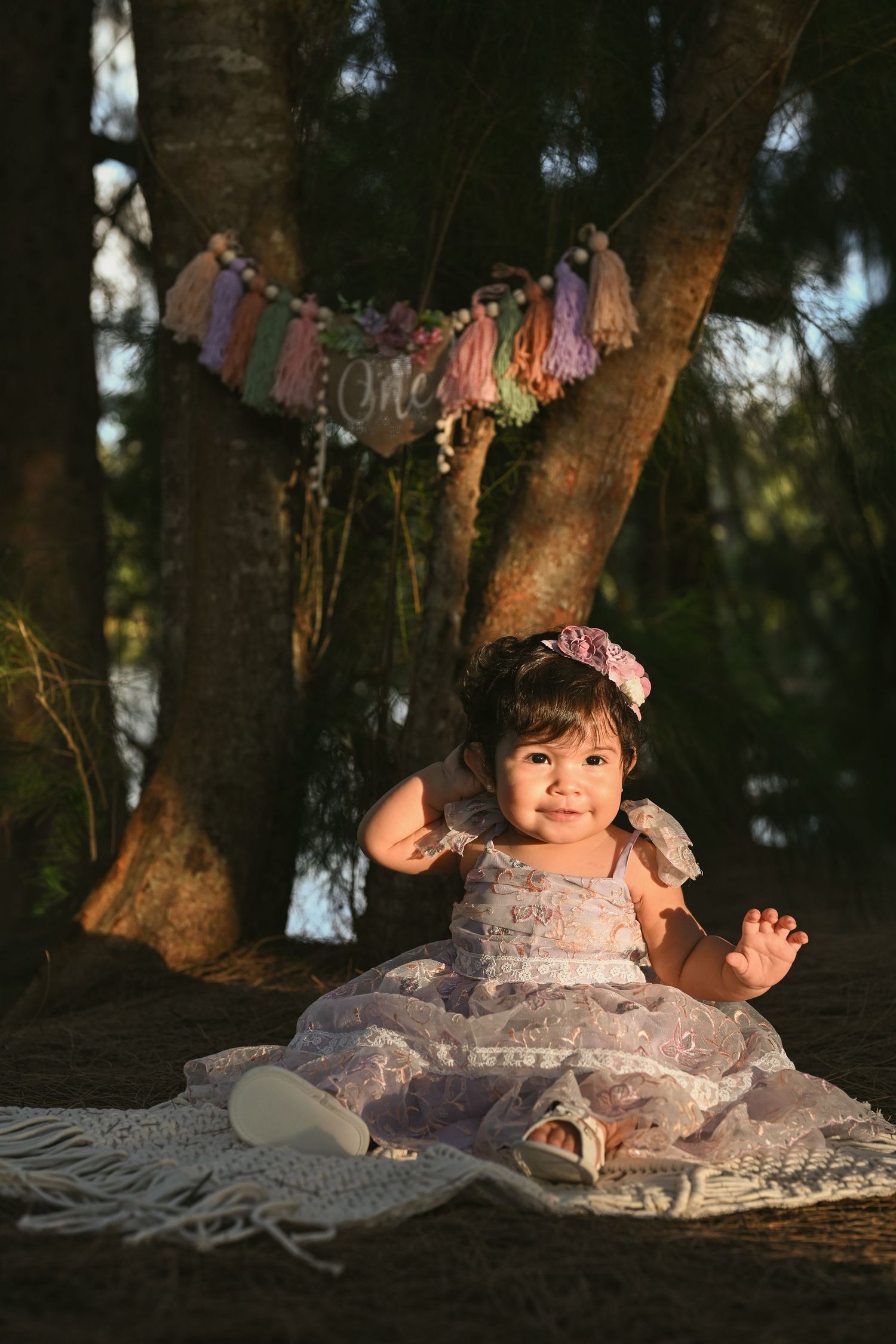 Baby in lace dress sits under decorative banners during outdoor photo session.