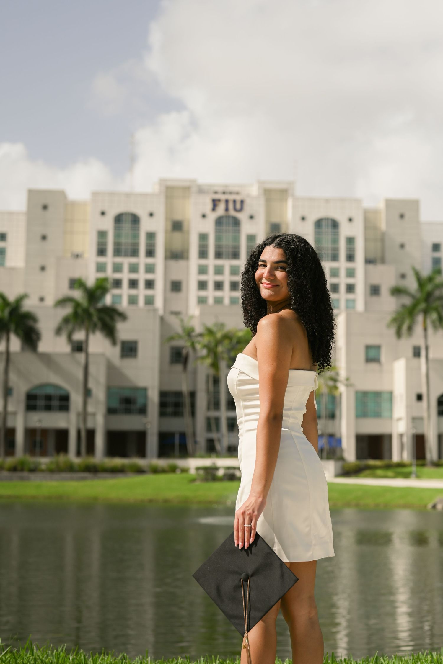White dress graduation photoshoot by a lake with palm trees and university buildings in background.