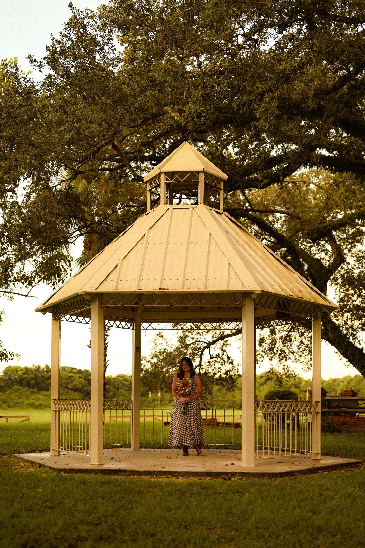 White wooden gazebo stands in a grassy field during golden hour with trees in the background.