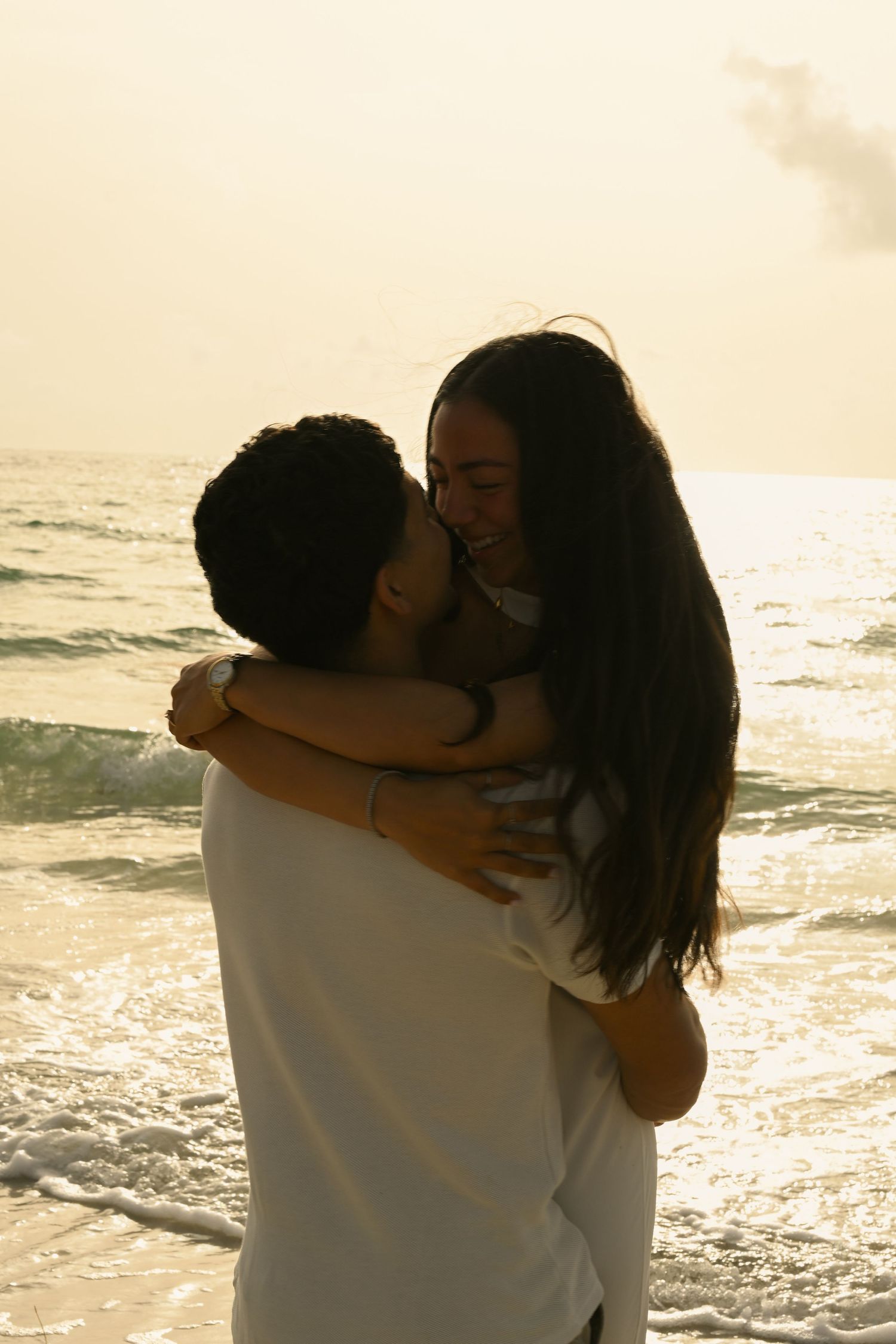 A romantic couple embraces on the beach during sunset with ocean waves in the background.