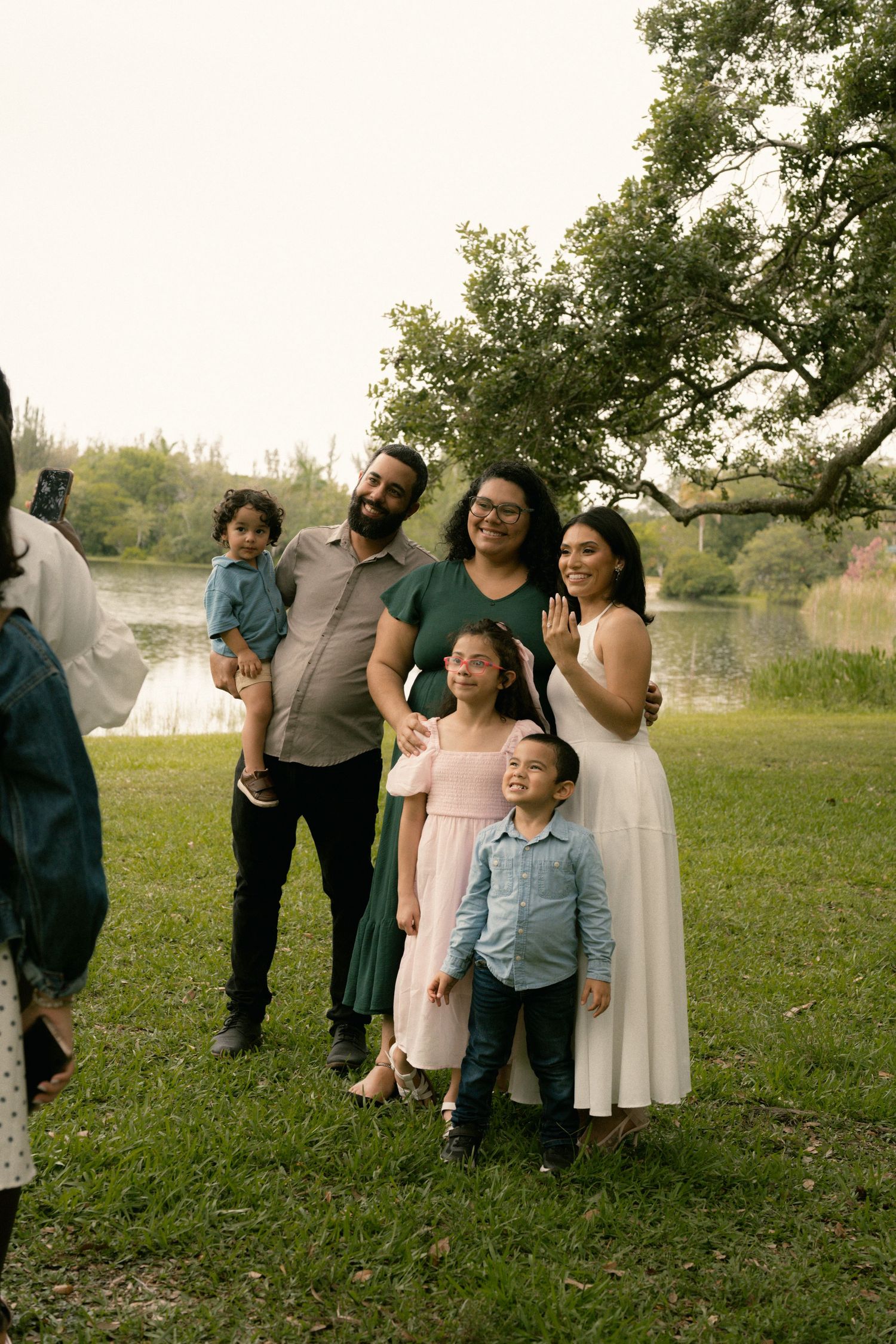 Happy family of five poses outdoors near a lake, parents with three children in semi-formal attire among green trees.