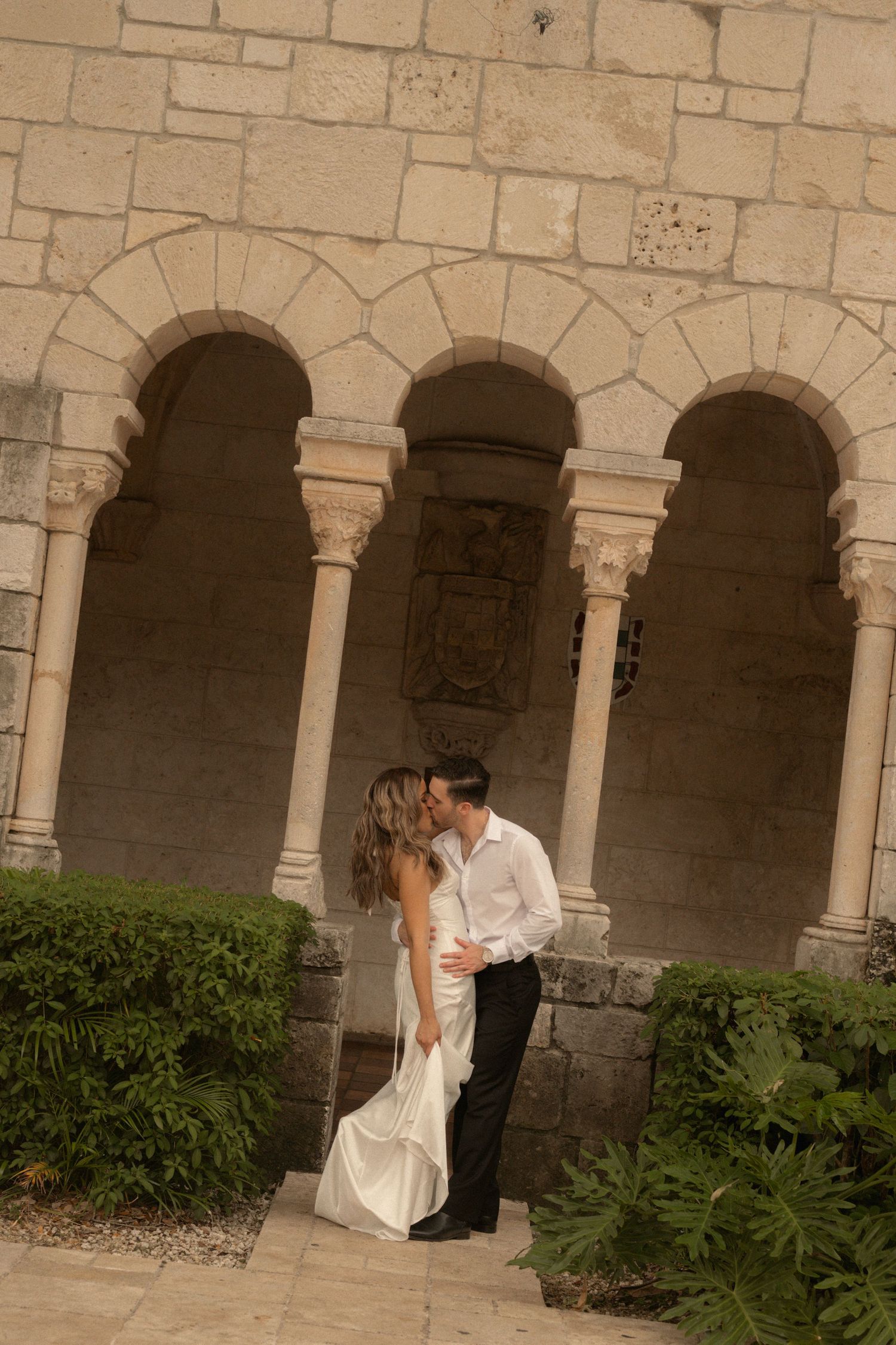 Couple in wedding attire kiss beneath ancient stone arches at a historic monastery, surrounded by lush green tropical plants.
