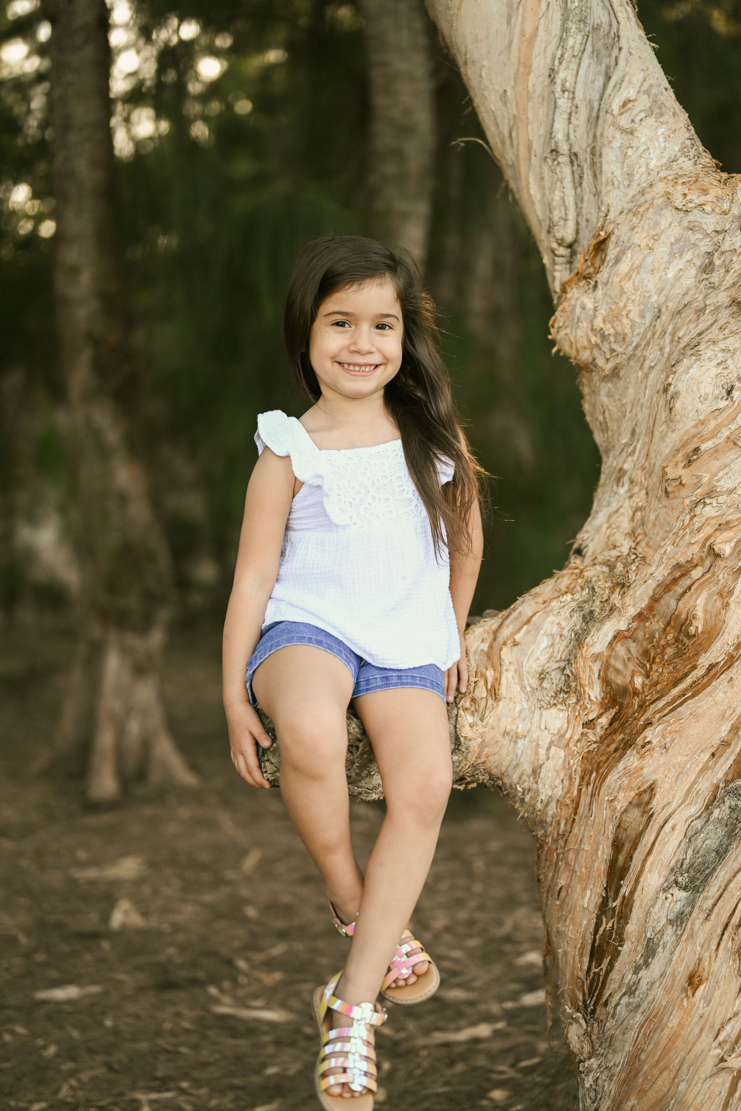 Young child in white top and blue shorts plays on a tree trunk in a wooded setting.