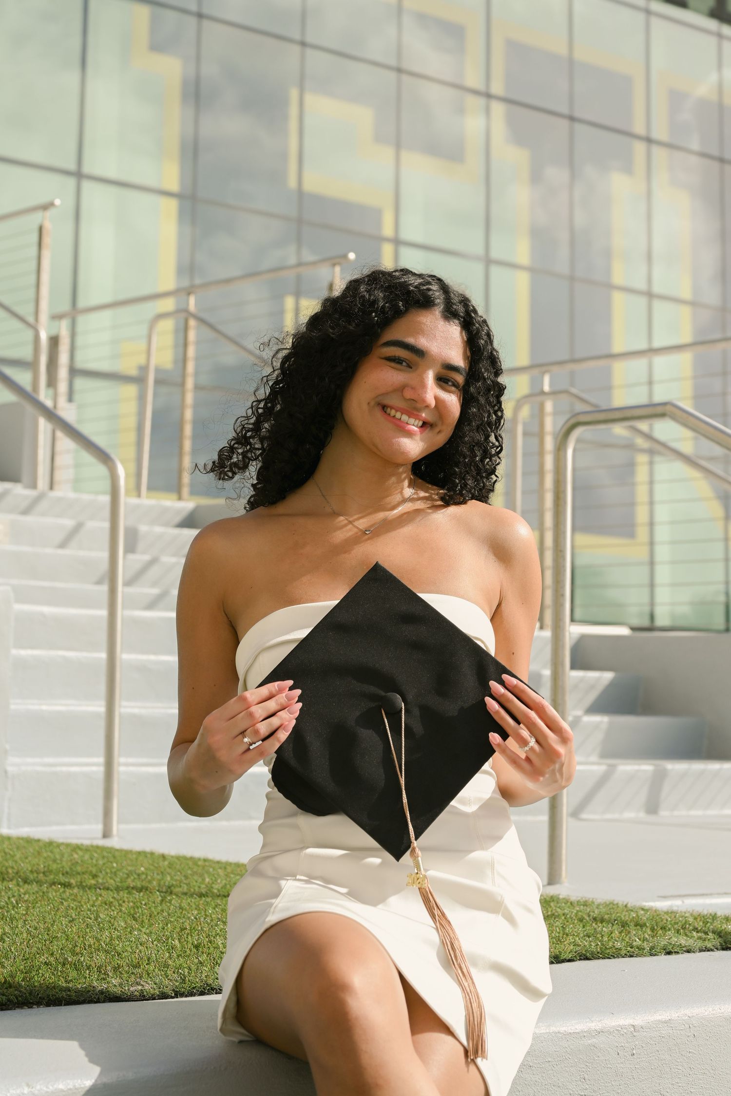 Graduate in white dress poses on steps with a black graduation cap in hand against modern building backdrop.