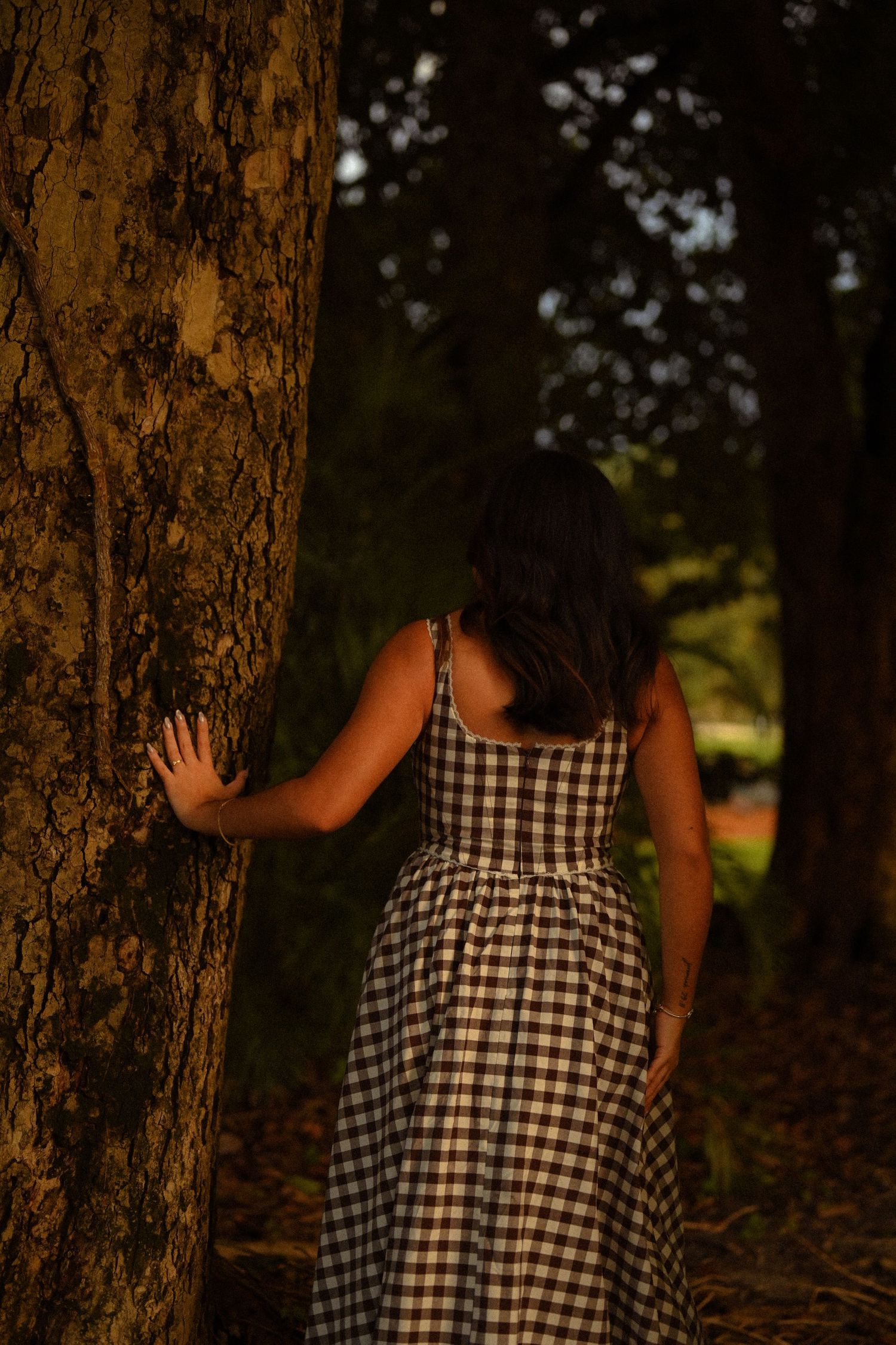 Someone in a gingham dress explores a grove of tall trees during golden hour.