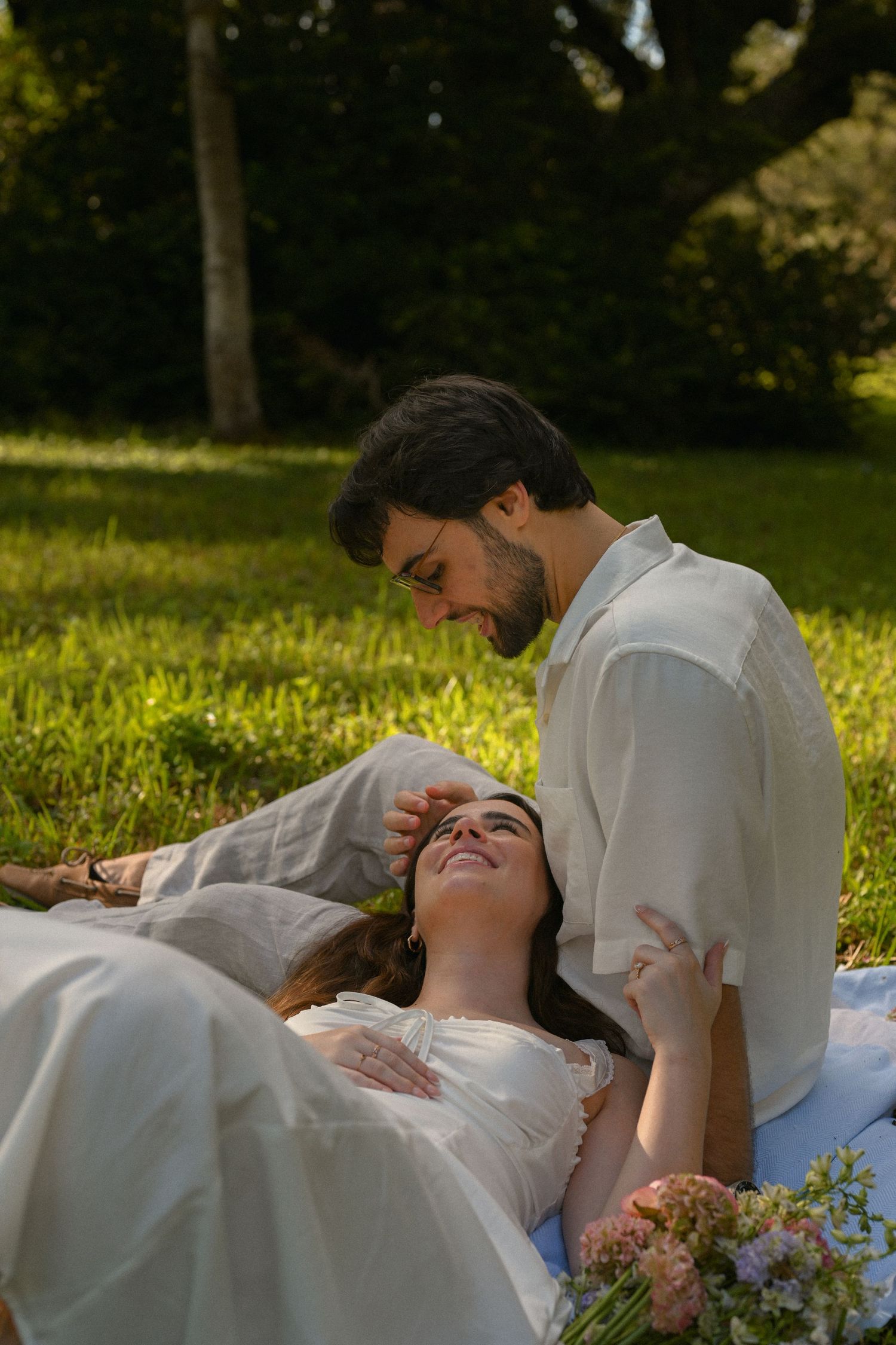 A couple relaxes together on a white blanket in a sunny grass field on a warm afternoon.