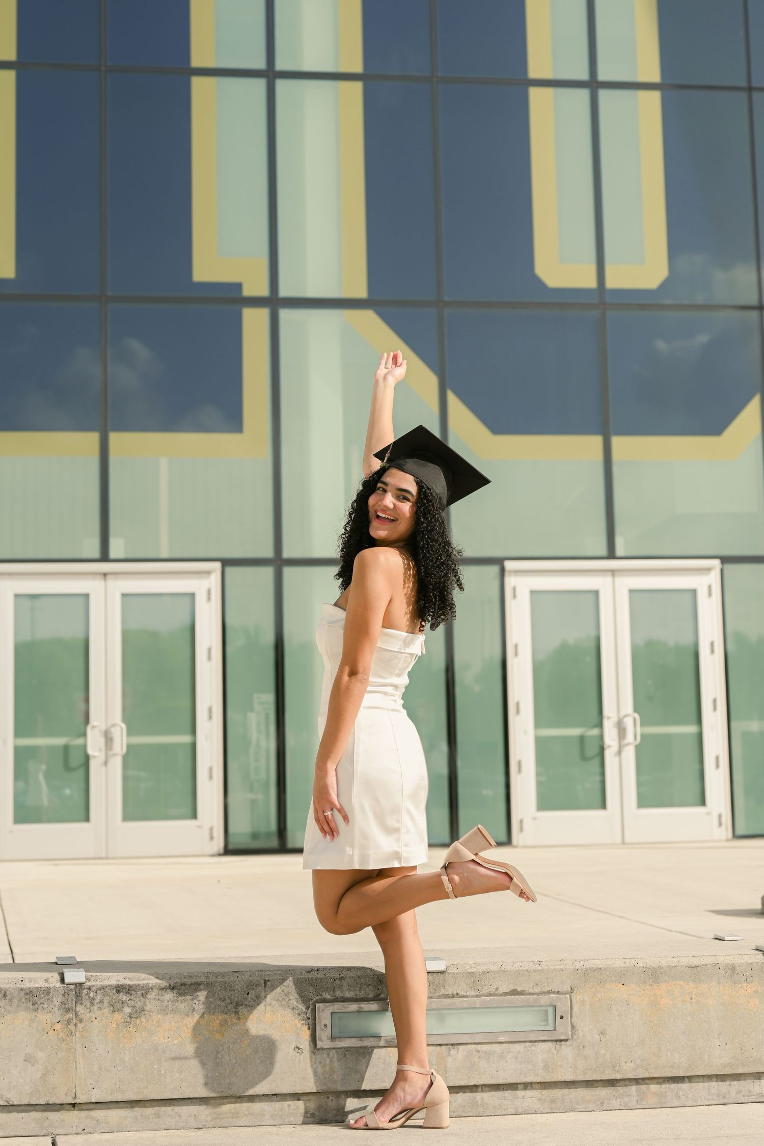 Graduate in cap and gown poses joyfully outside modern building entrance.