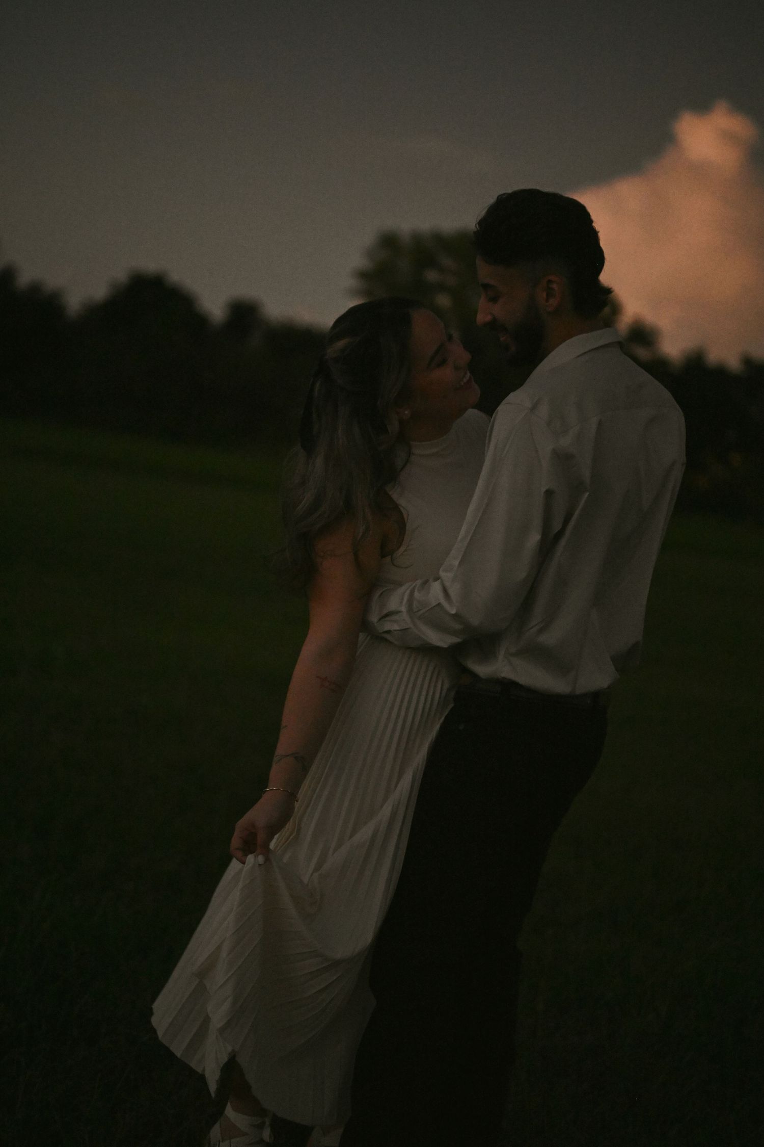 A romantic couple embraces in a field during twilight hours.