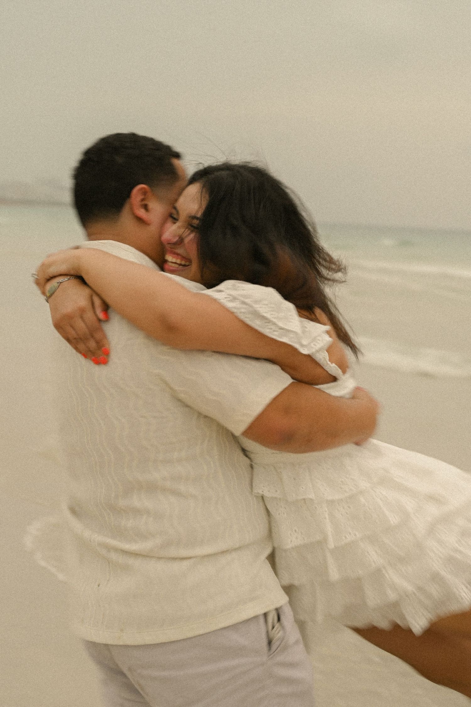 A couple in white embraces tenderly on the beach with ocean waves in the background.