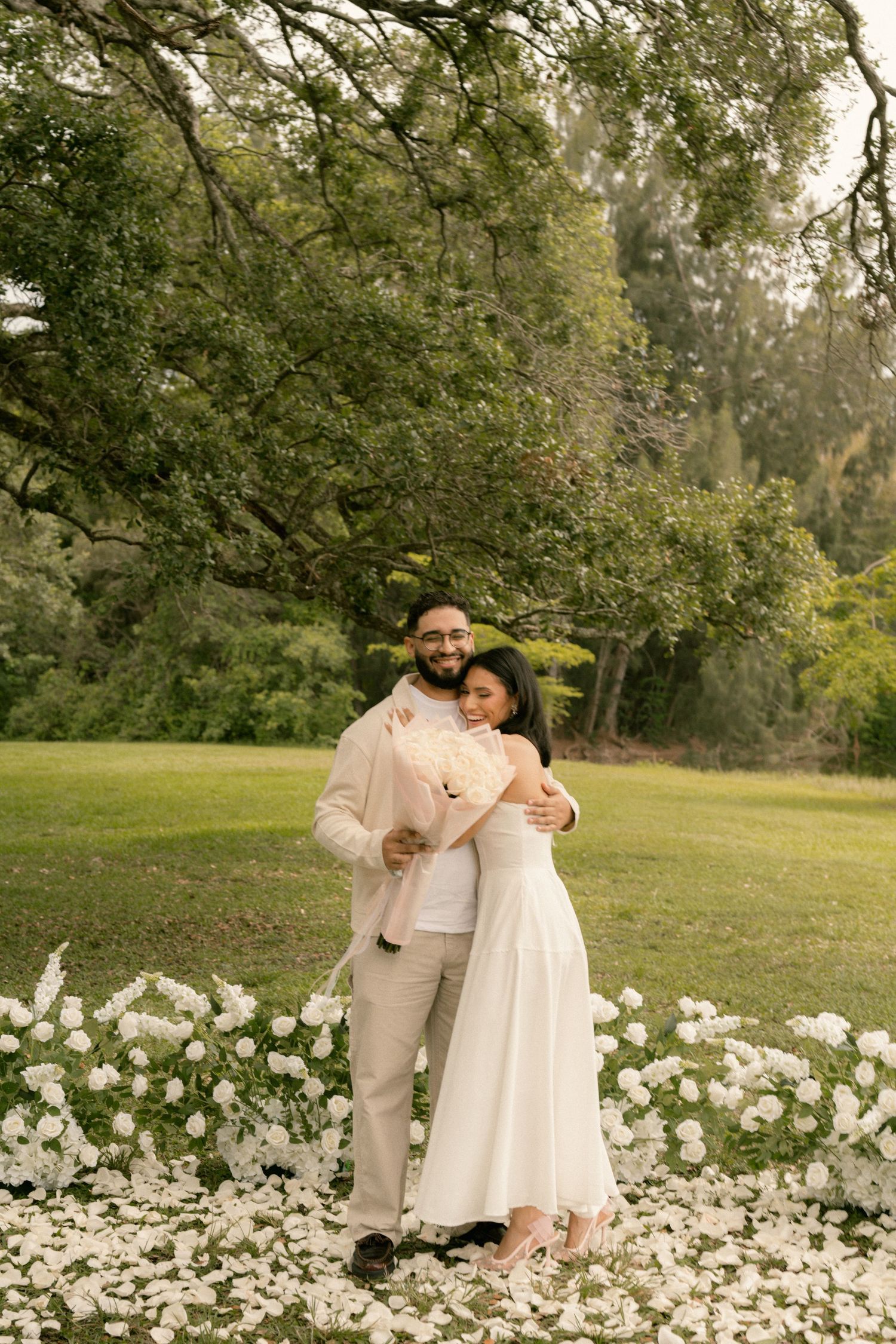 Couple embracing in a lush garden setting, bride in white midi dress holding bouquet, groom in beige suit, white flowers at feet.