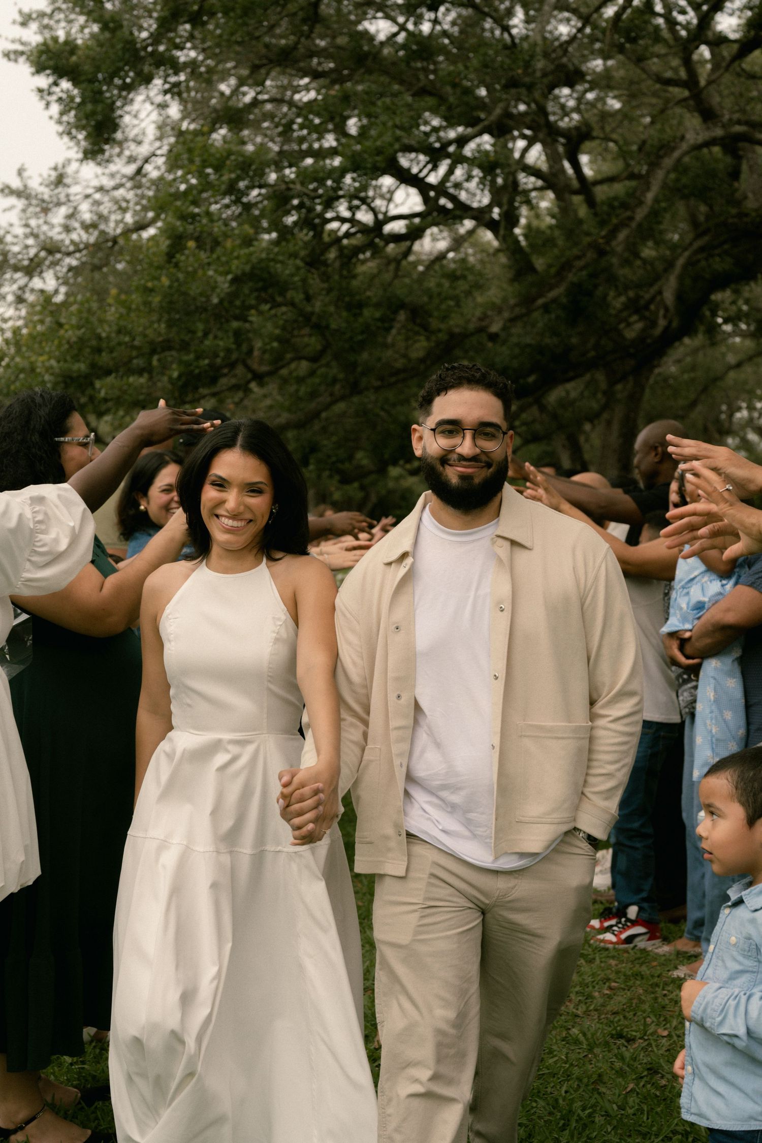 Beaming newlyweds walk hand-in-hand through applauding guests outdoors, bride in white halter dress, groom in beige jacket.