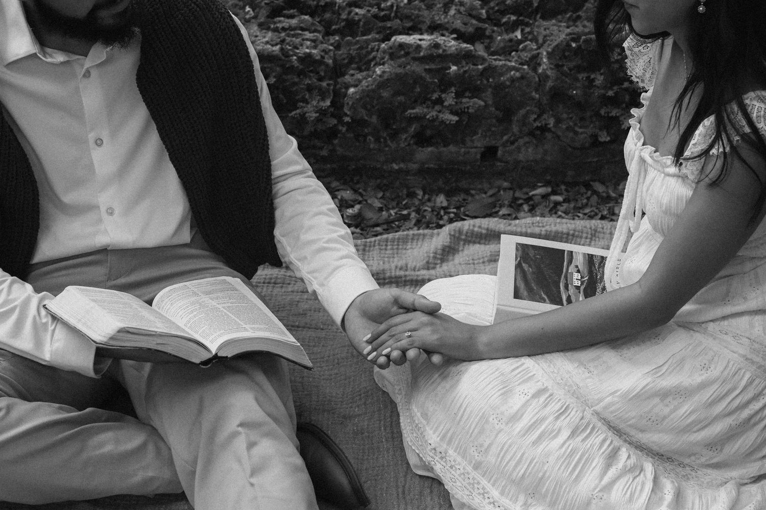 A couple sits together on a bench holding hands while one reads from an open book in black and white.