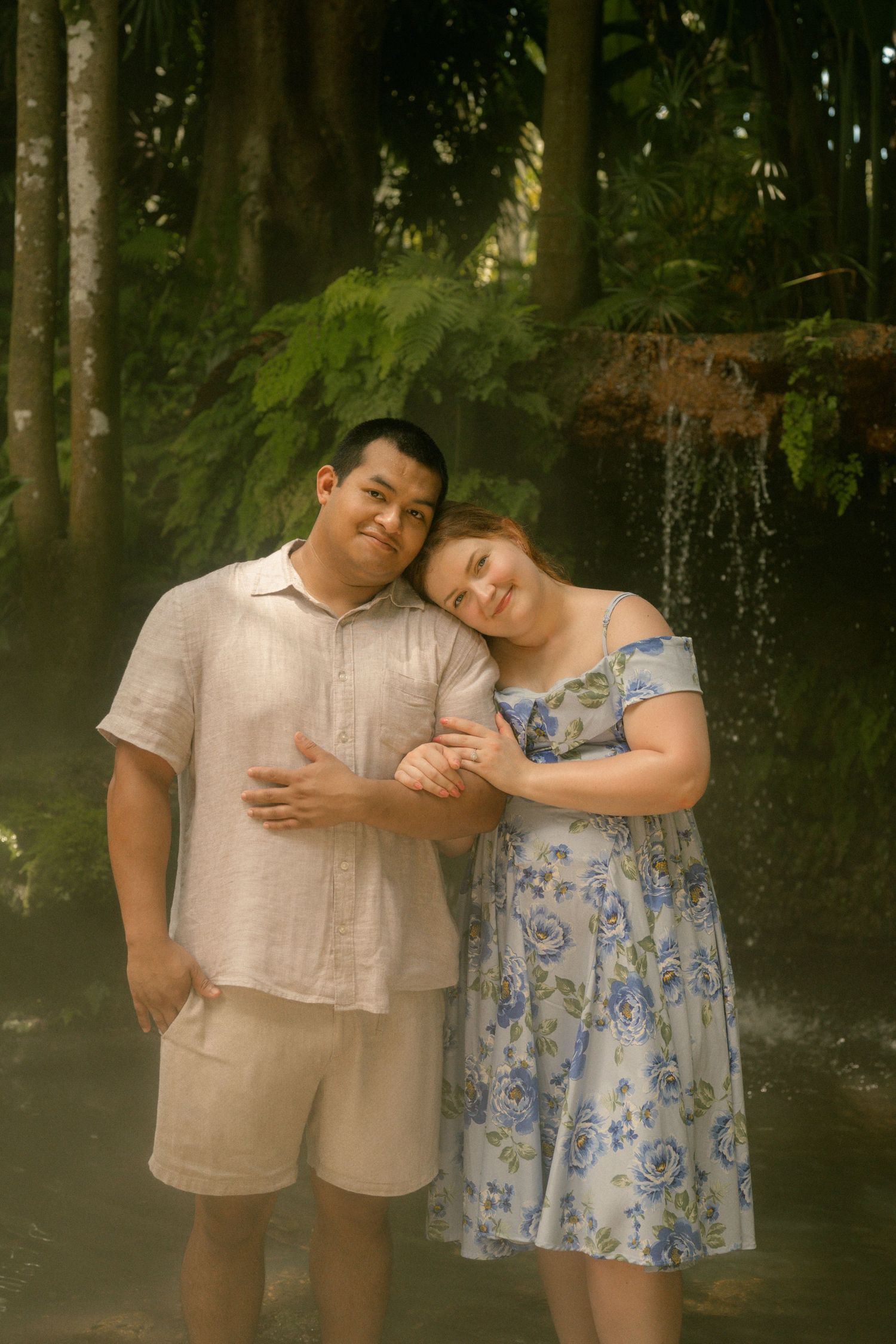 Happy couple posing together in a lush tropical garden, woman in floral dress leaning on man's shoulder.