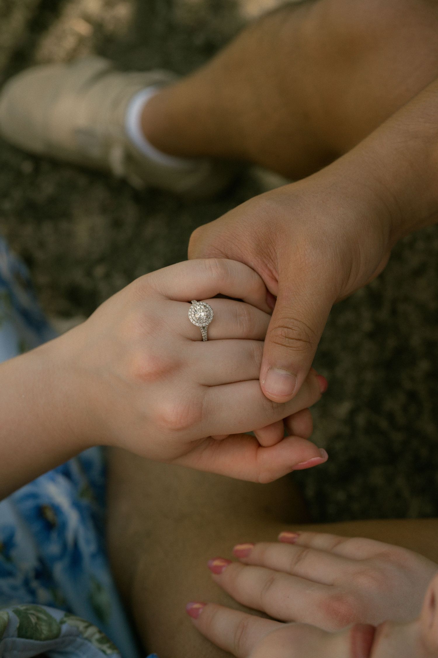 Close-up of a couple holding hands showing a round halo diamond engagement ring on the woman's finger.