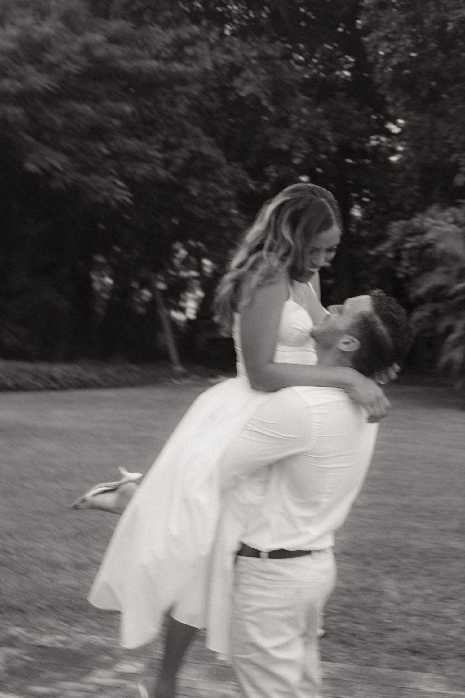 Black and white photo of a man lifting a smiling woman in a white dress in a joyful embrace on a grassy lawn surrounded by trees.