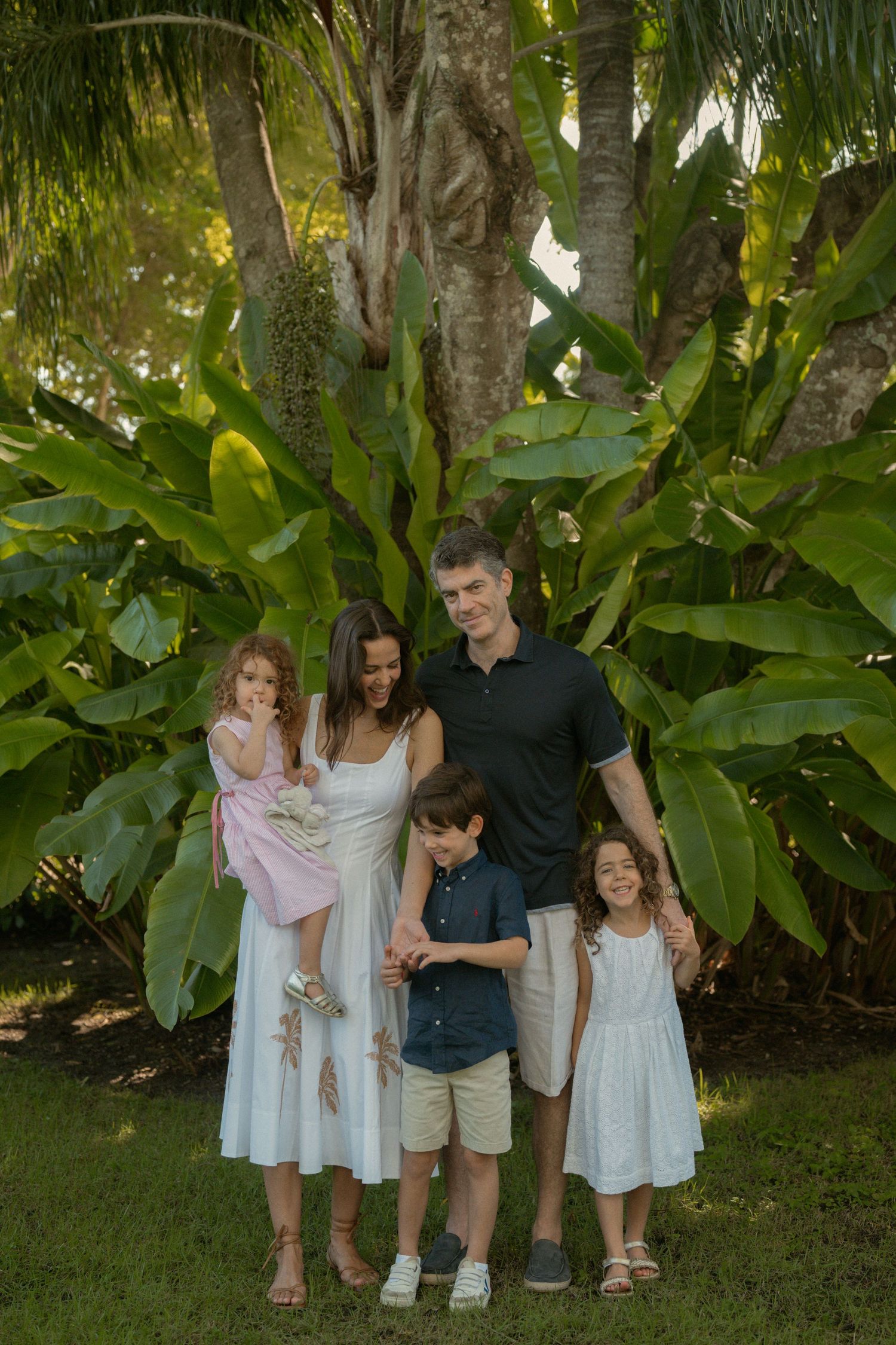 Family of five in coordinated white and navy outfits posing together on a green lawn surrounded by lush tropical foliage and palm trees.