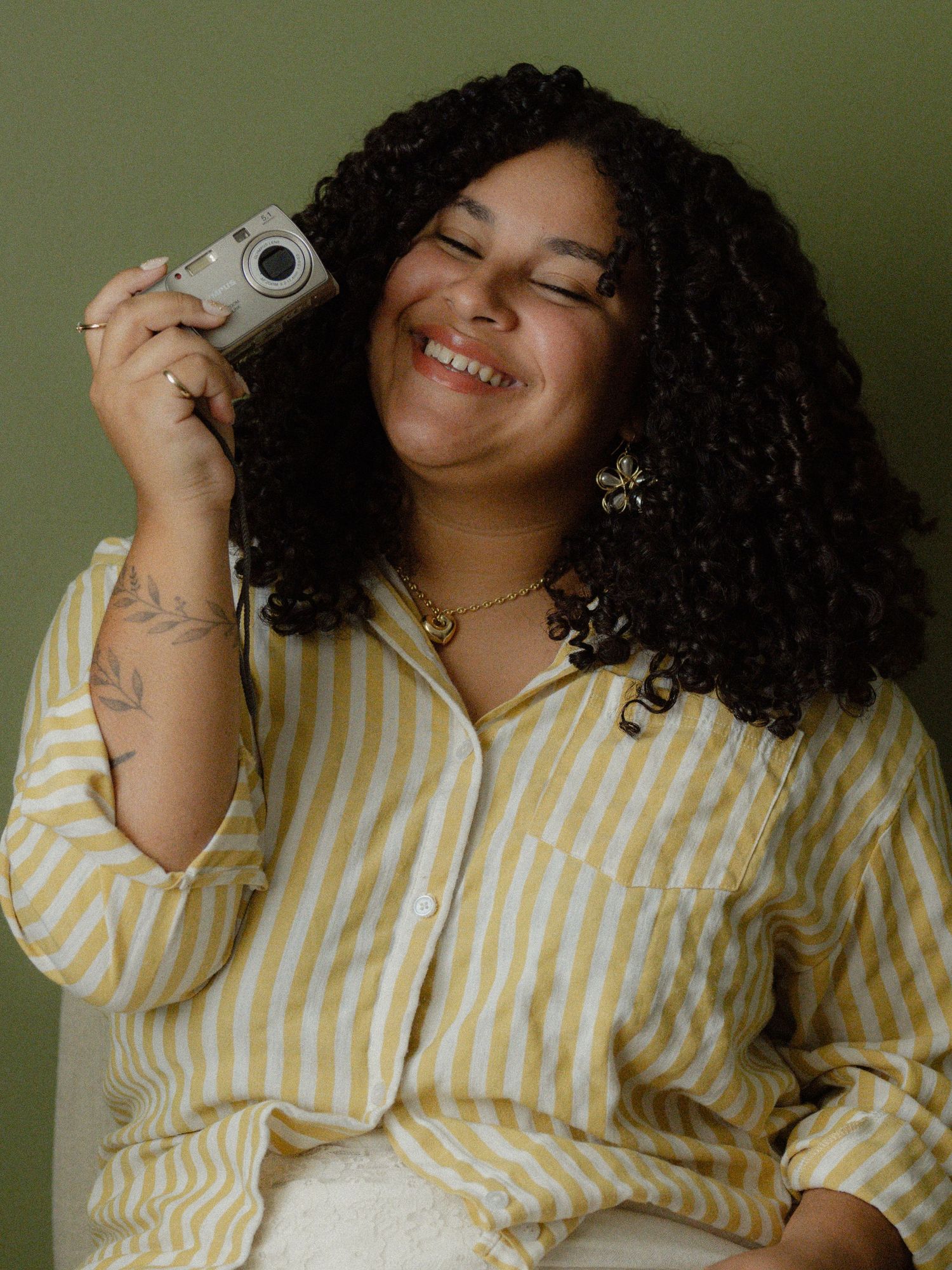 Smiling woman with curly hair holds a vintage camera, wearing a yellow striped shirt and gold jewelry.