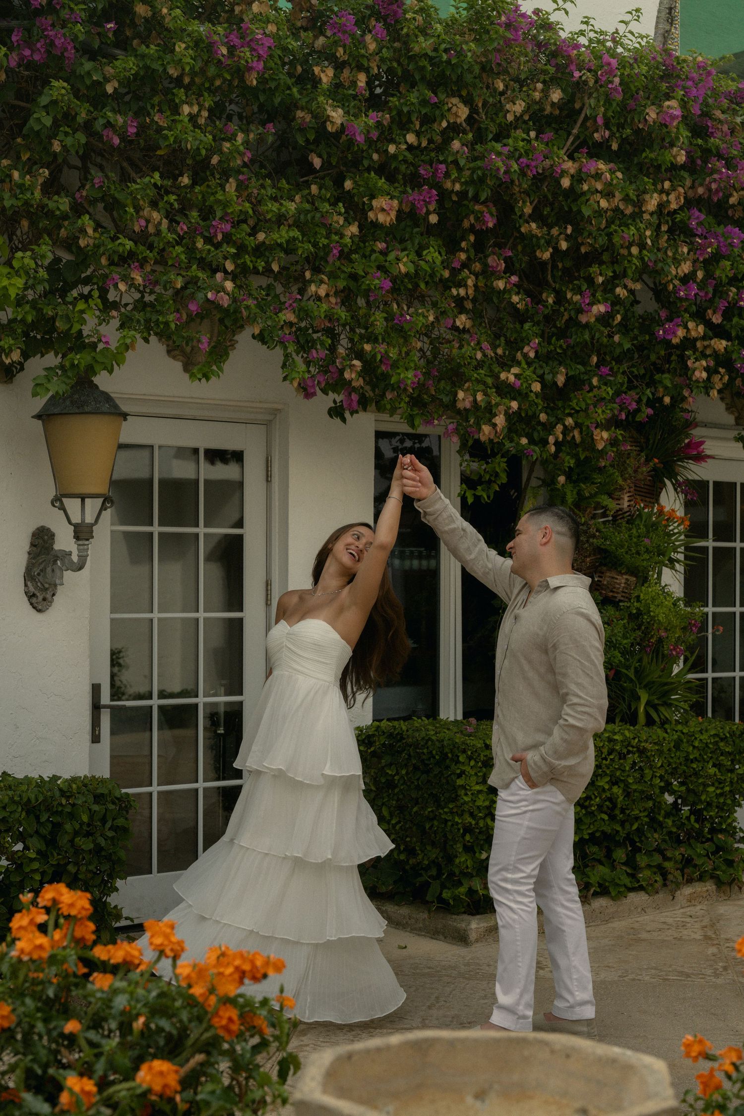 Couple dancing outdoors in white attire under blooming bougainvillea with orange flowers and white building backdrop.