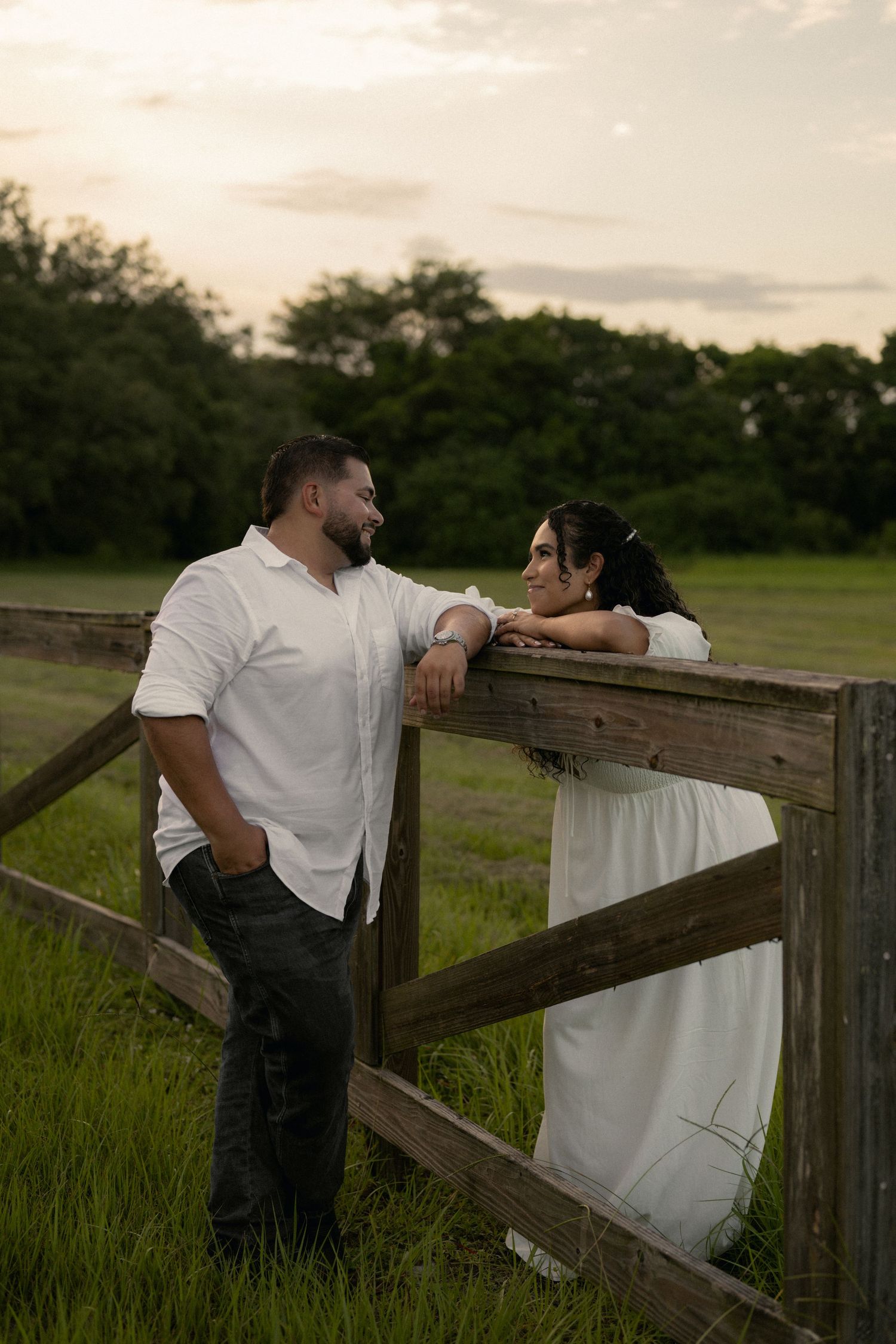 Couple in white attire sharing a romantic moment by a rustic wooden fence in a lush green field at sunset.