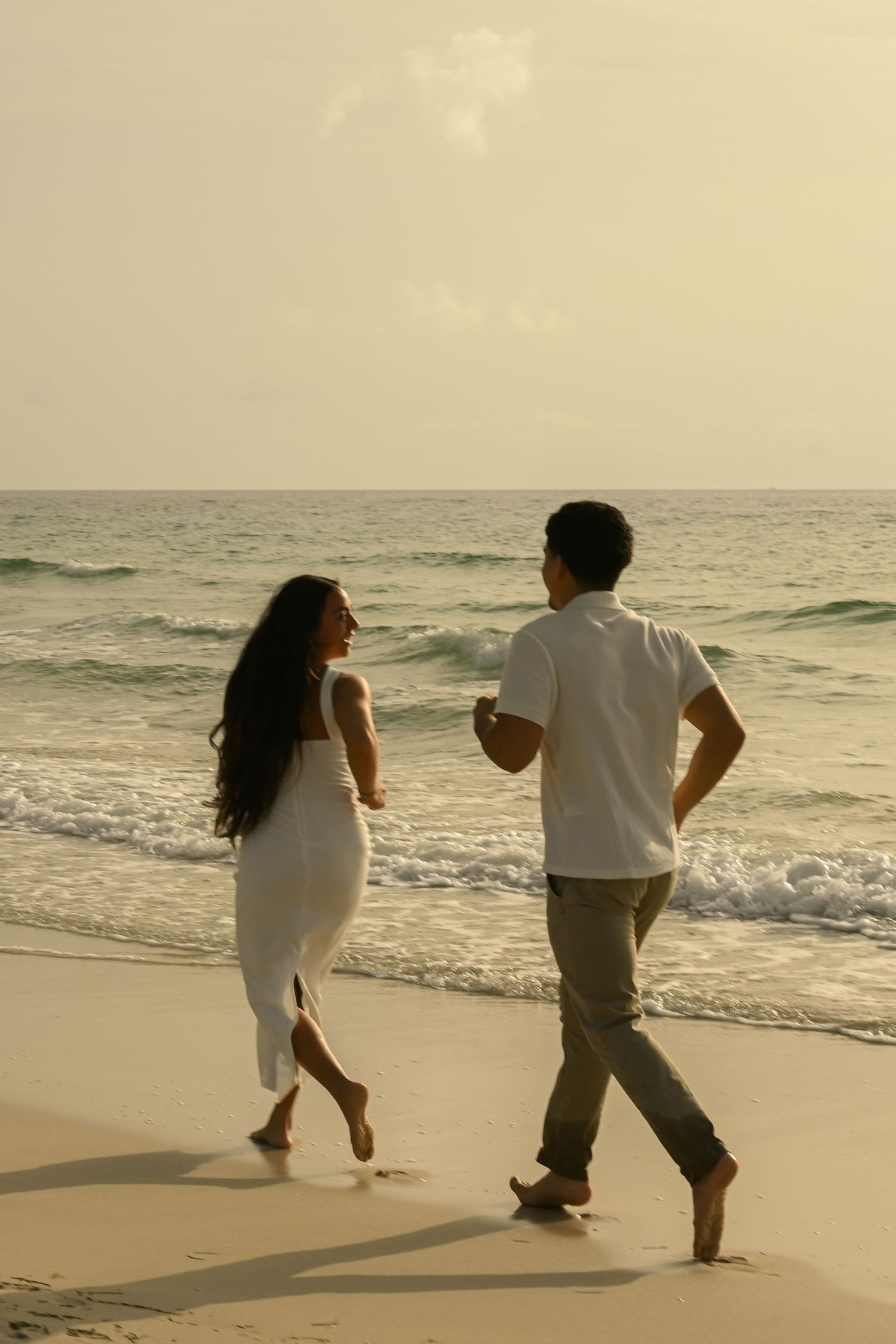 A couple runs playfully along a sunset beach with waves crashing in the background.