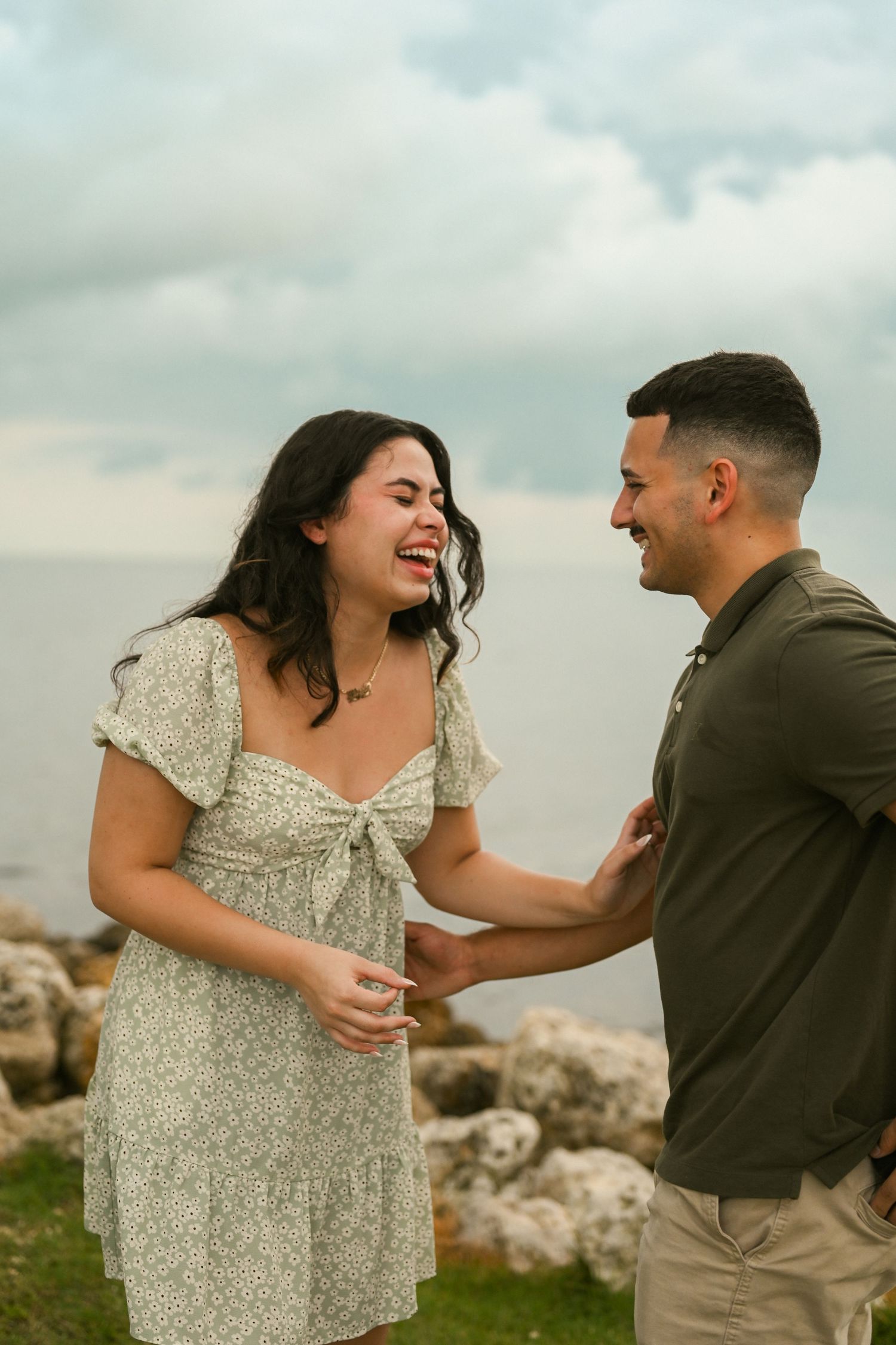 A couple shares joyful laughter together during a romantic photoshoot on a rocky coastline at sunset.