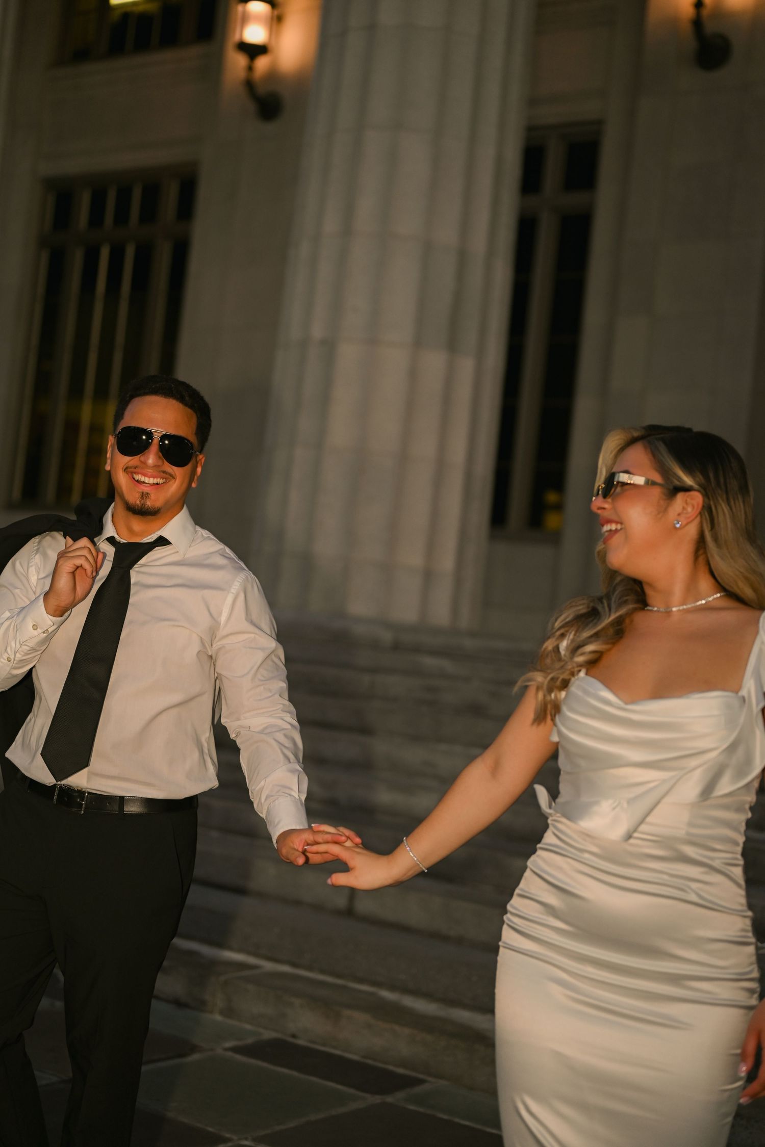 Couple in wedding attire dancing and laughing together outside a classical building.