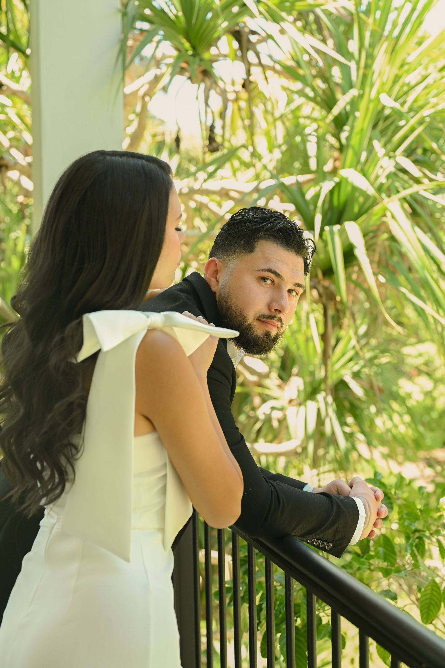 A couple shares tender moments on a balcony with tropical plants in the background.