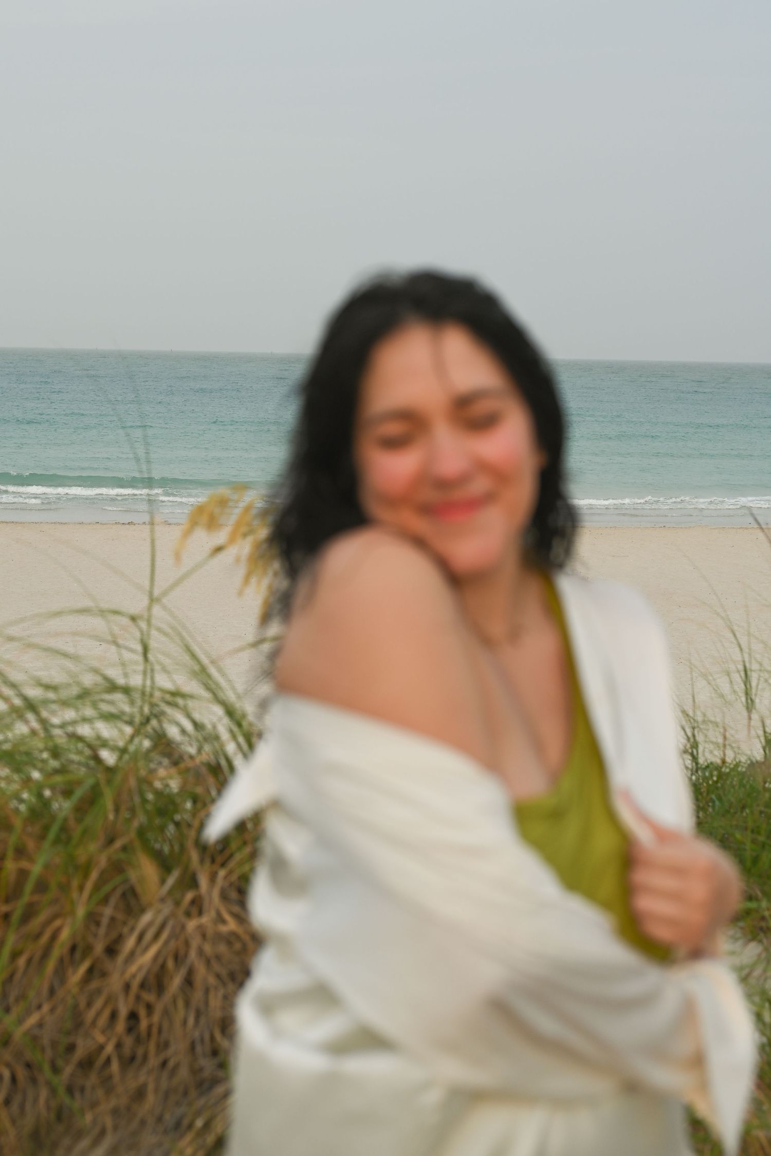 Person in white wrap stands near beach grass and ocean.