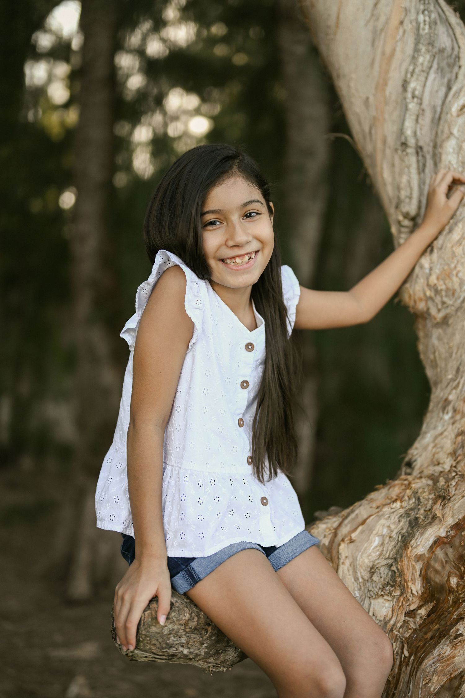 A child in white clothing plays on a tree branch in a wooded setting.
