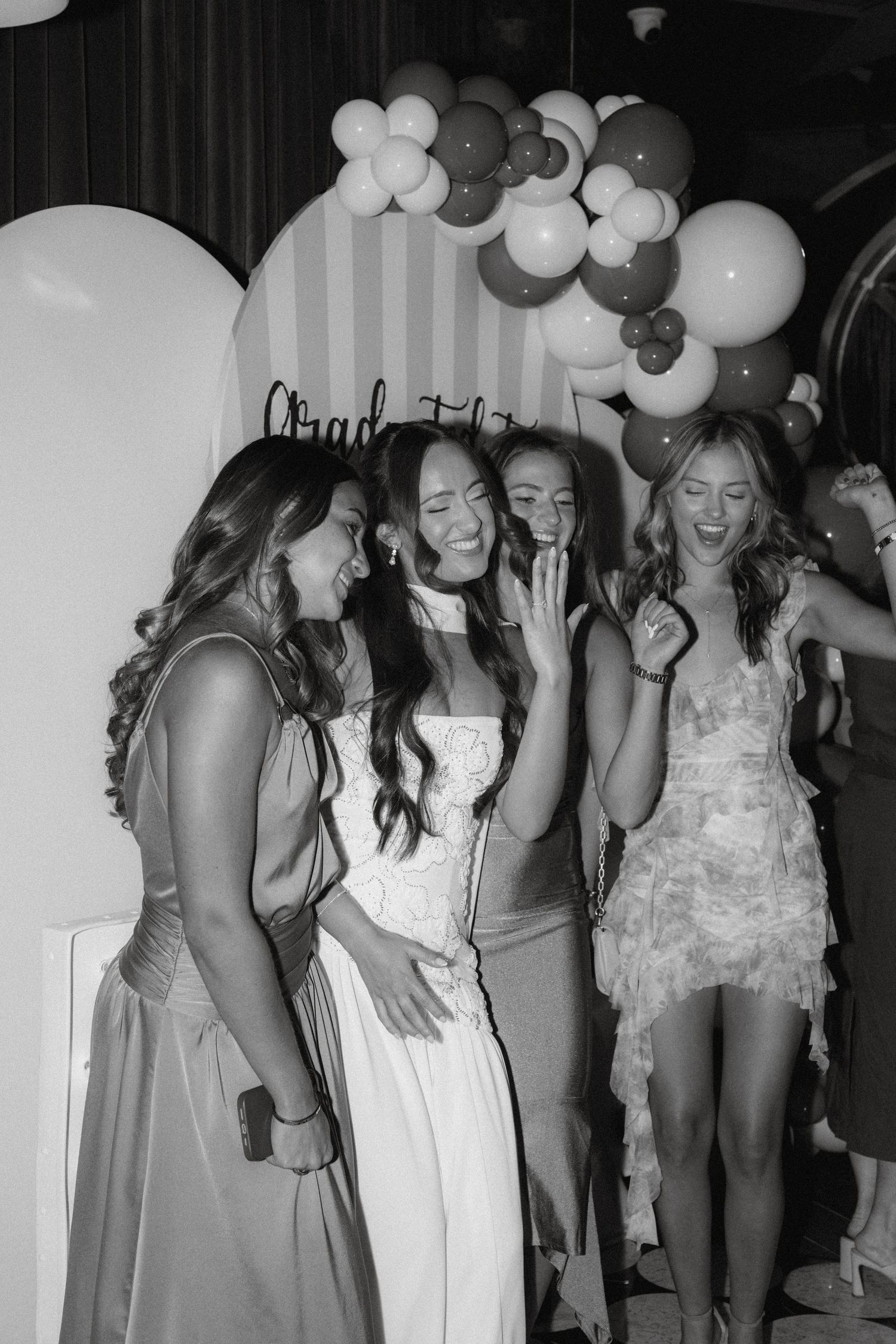 Four young women laughing and celebrating at a graduation party with balloon arch backdrop in black and white photo.