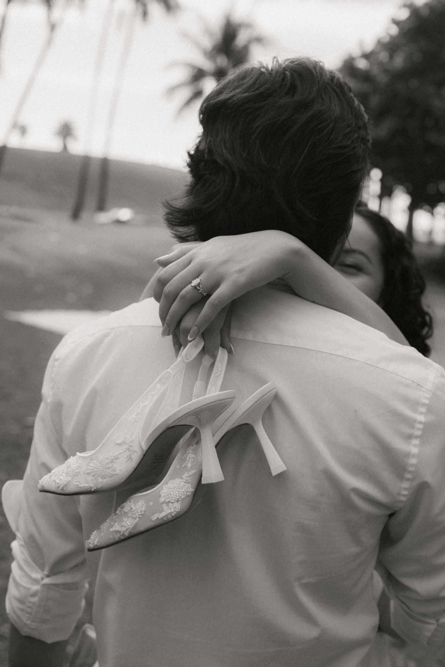 Black and white photo of a bride holding heels while embracing groom outdoors near palm trees.