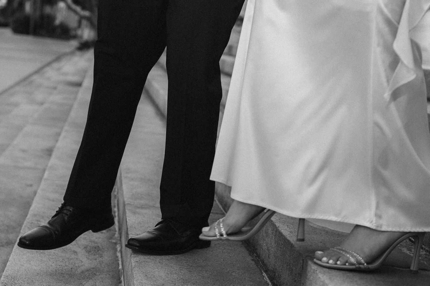 Black and white photo of bride and groom feet on steps, wedding dress and dress shoes visible.