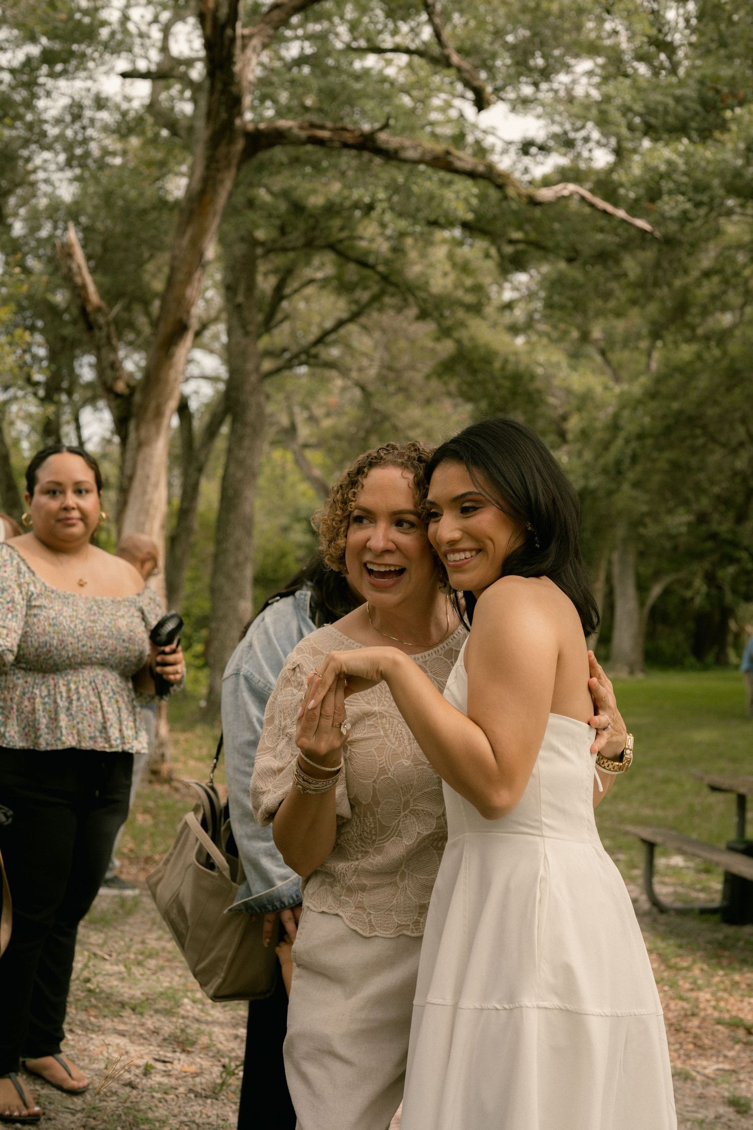 Two women embrace and smile outdoors at a wedding celebration surrounded by trees.