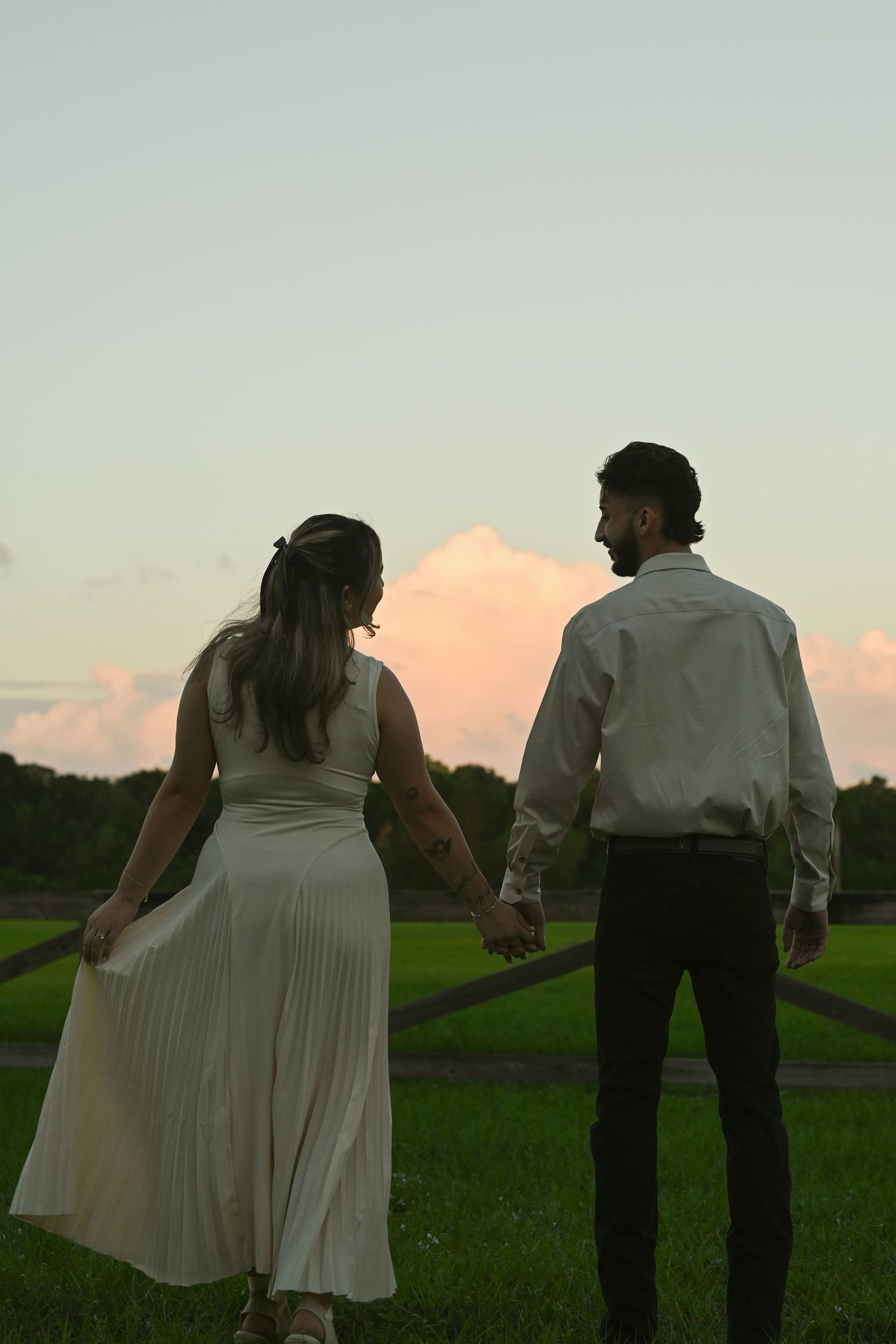 A couple in light colored clothing holds hands while walking in a grassy field during sunset.