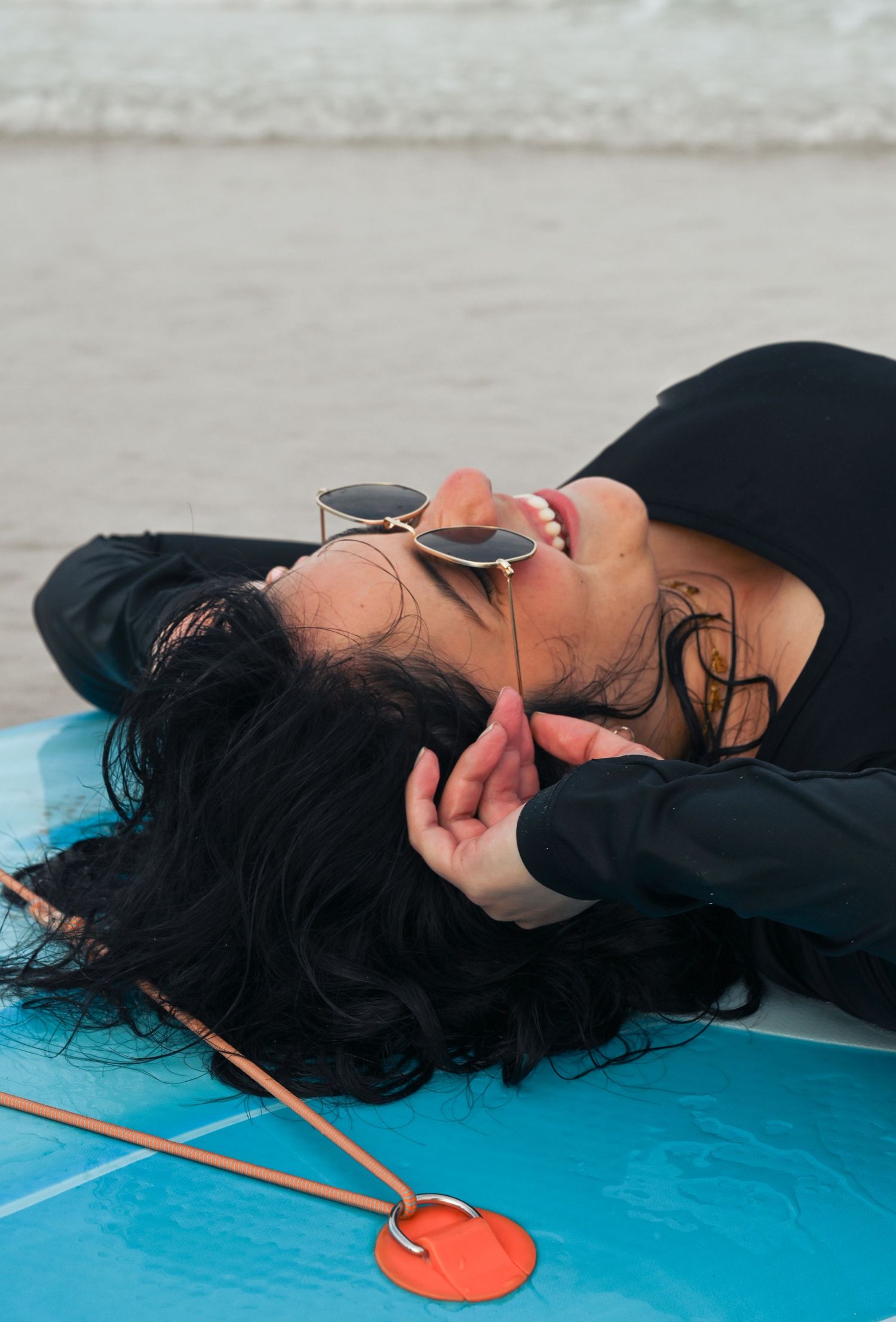 Person in black top relaxing on blue surfboard with sunglasses at beach.