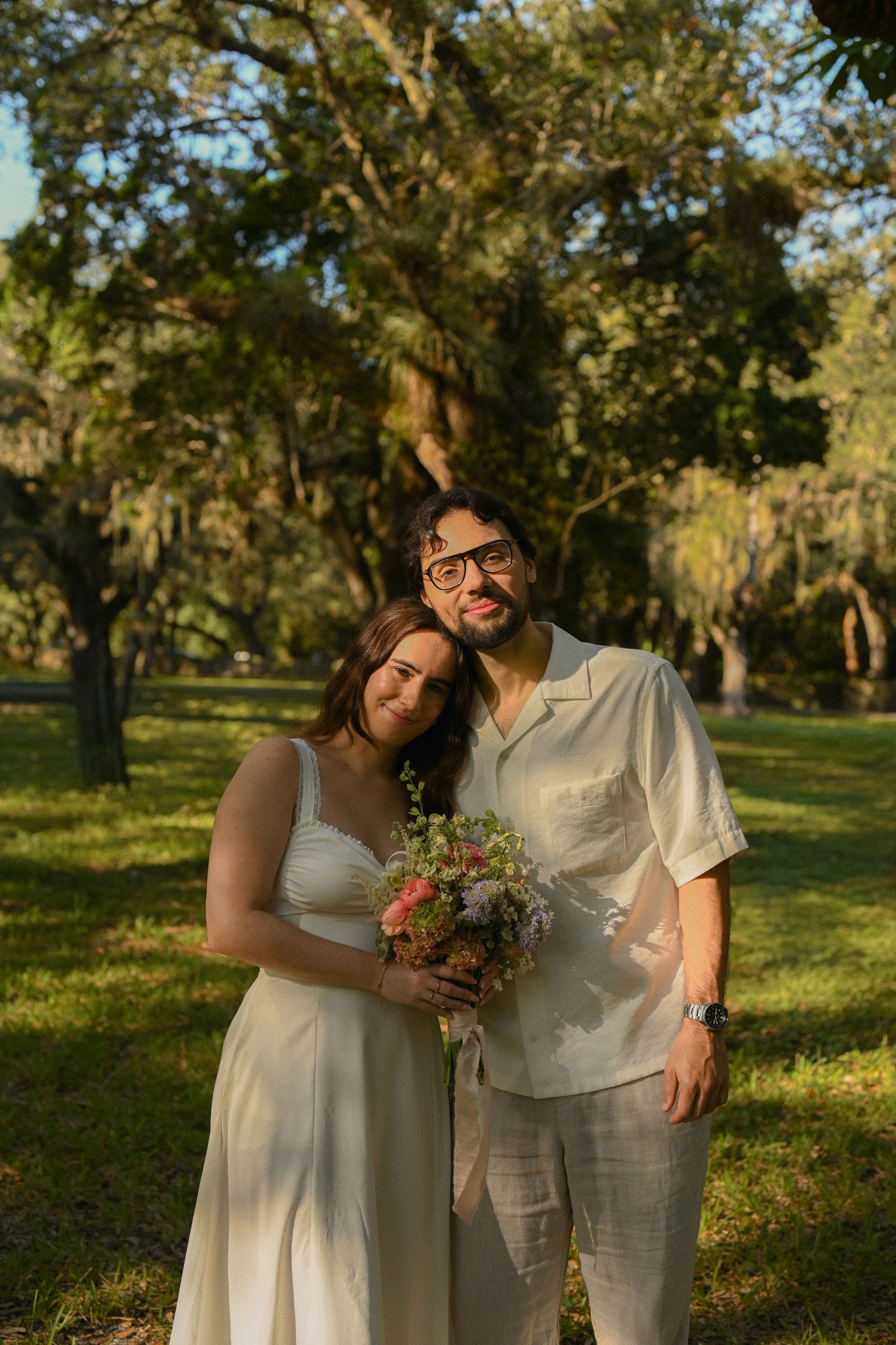 A couple in white outfits embrace and smile while standing in a sunlit park with trees in the background.