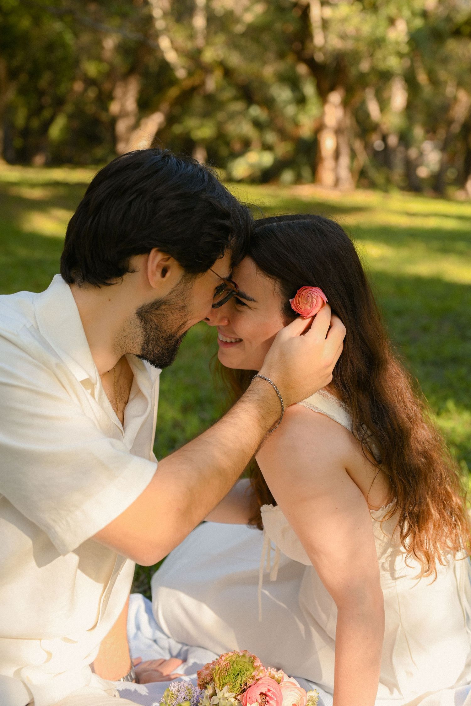 Romantic couple sitting in a sunny park, man gently touching woman's hair while holding a pink flower bouquet.