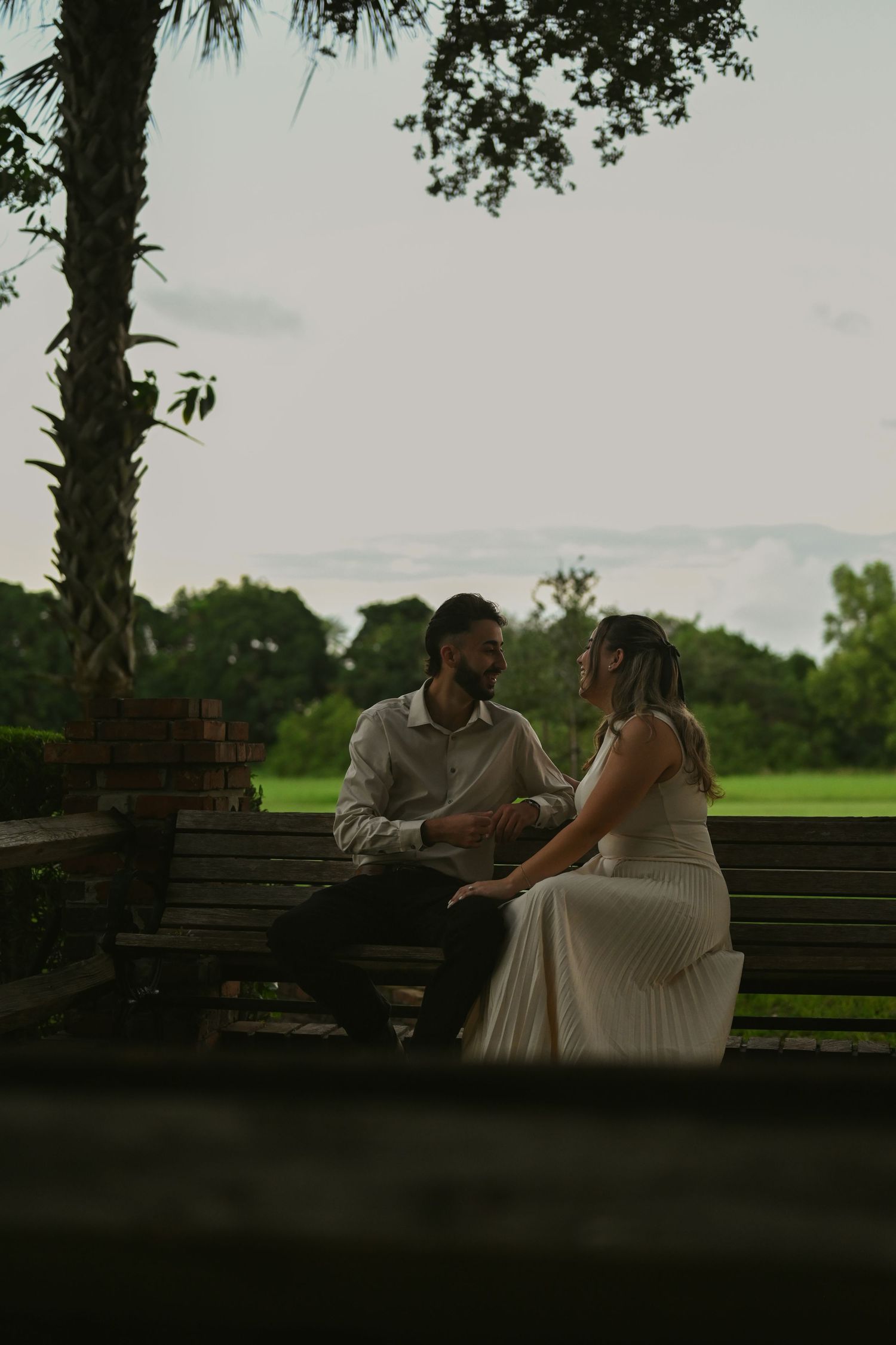Couple in wedding attire sitting on a bench surrounded by lush greenery, sharing an intimate moment.
