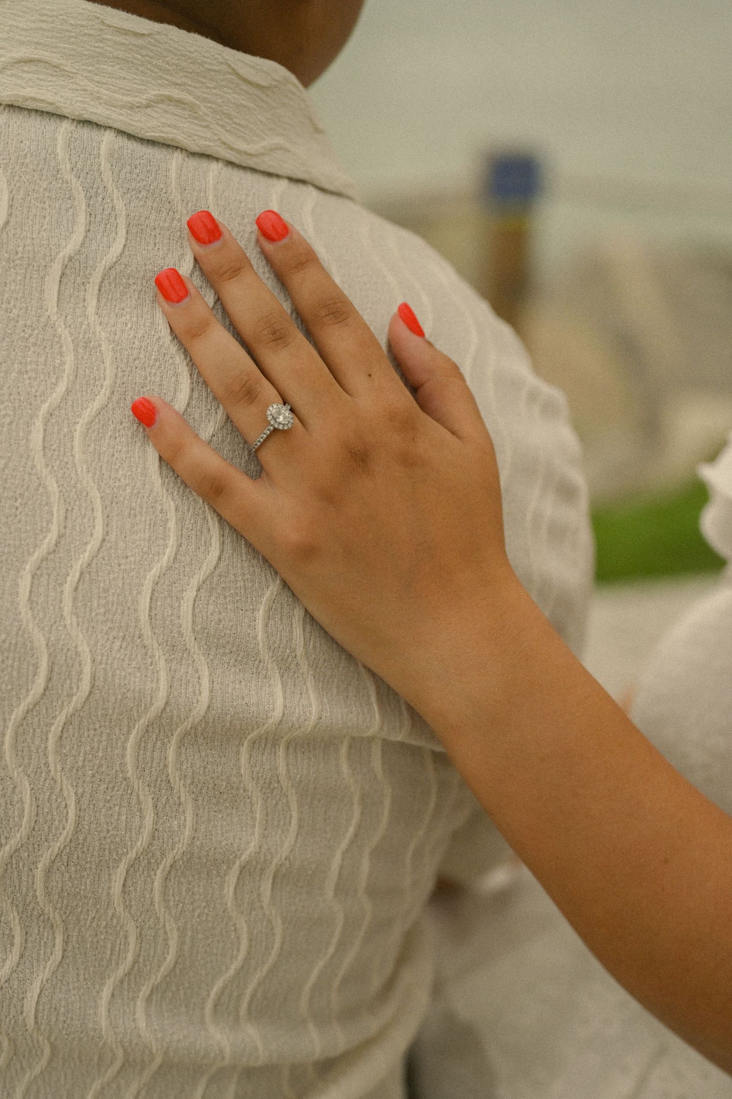 Close-up of a hand with bright red nails and engagement ring against white textured fabric.