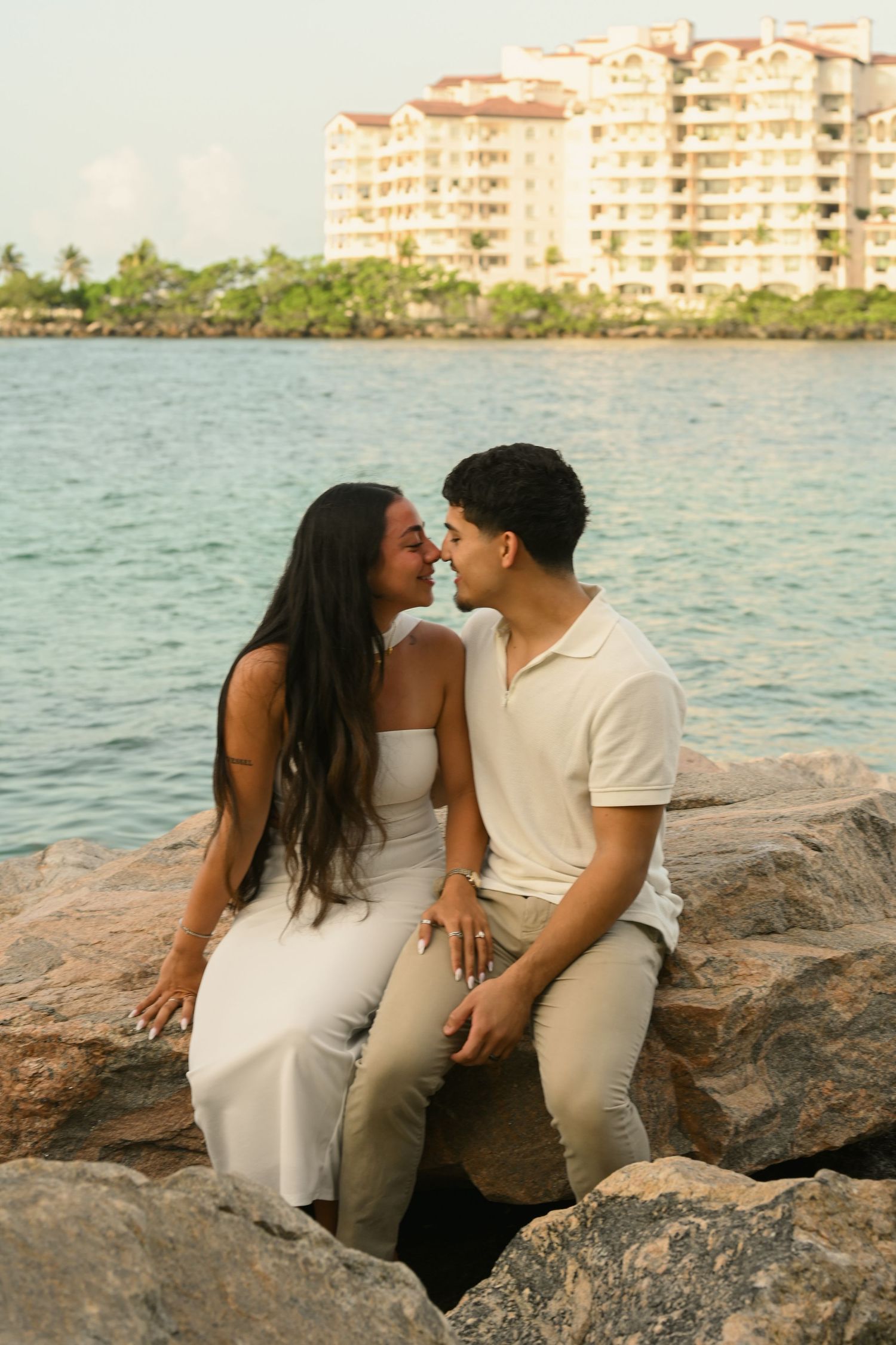 Couple in white outfits sitting close together on coastal rocks with waterfront buildings in background.