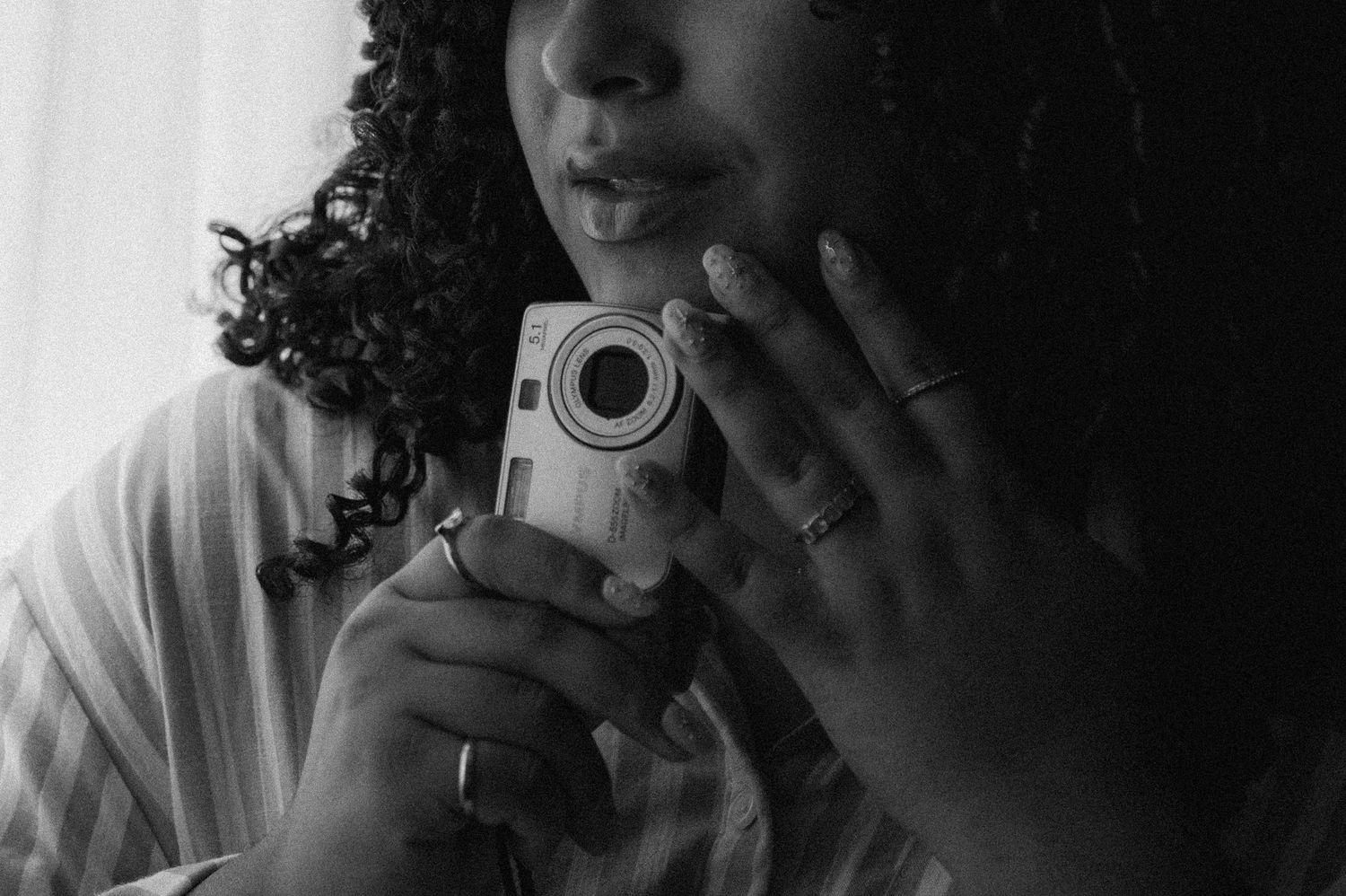 Black and white photo of a young woman with curly hair holding a small digital camera close to her face.