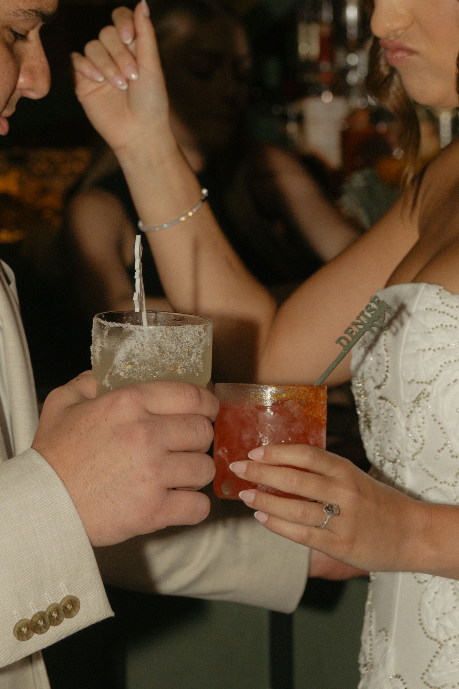 Bride and groom clinking cocktail glasses at wedding reception, bride wearing white lace dress with floral tattoo.