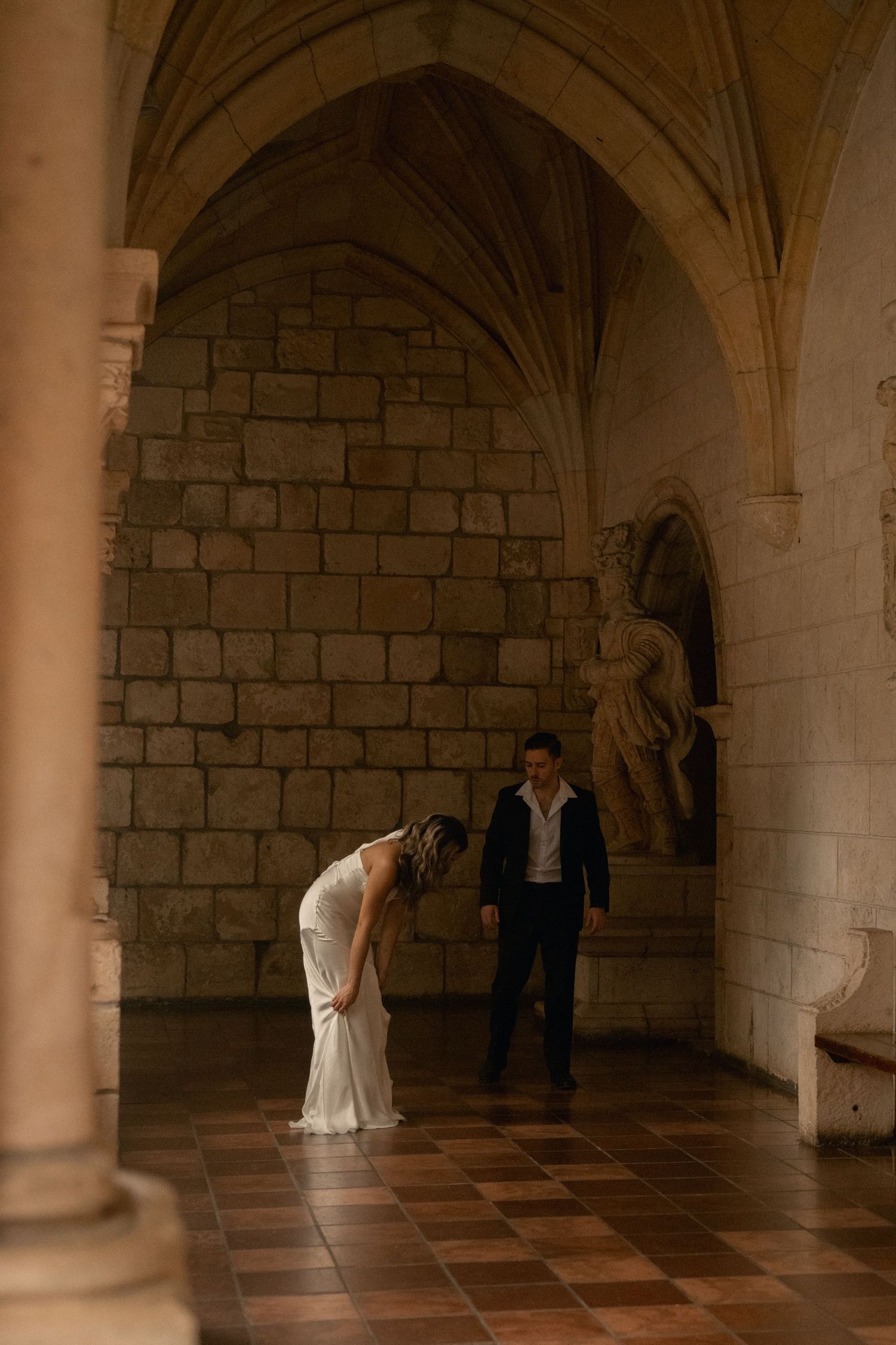 A bride in a white gown bends forward as a groom in a dark suit watches in a stone cloister with vaulted arches and a statue.