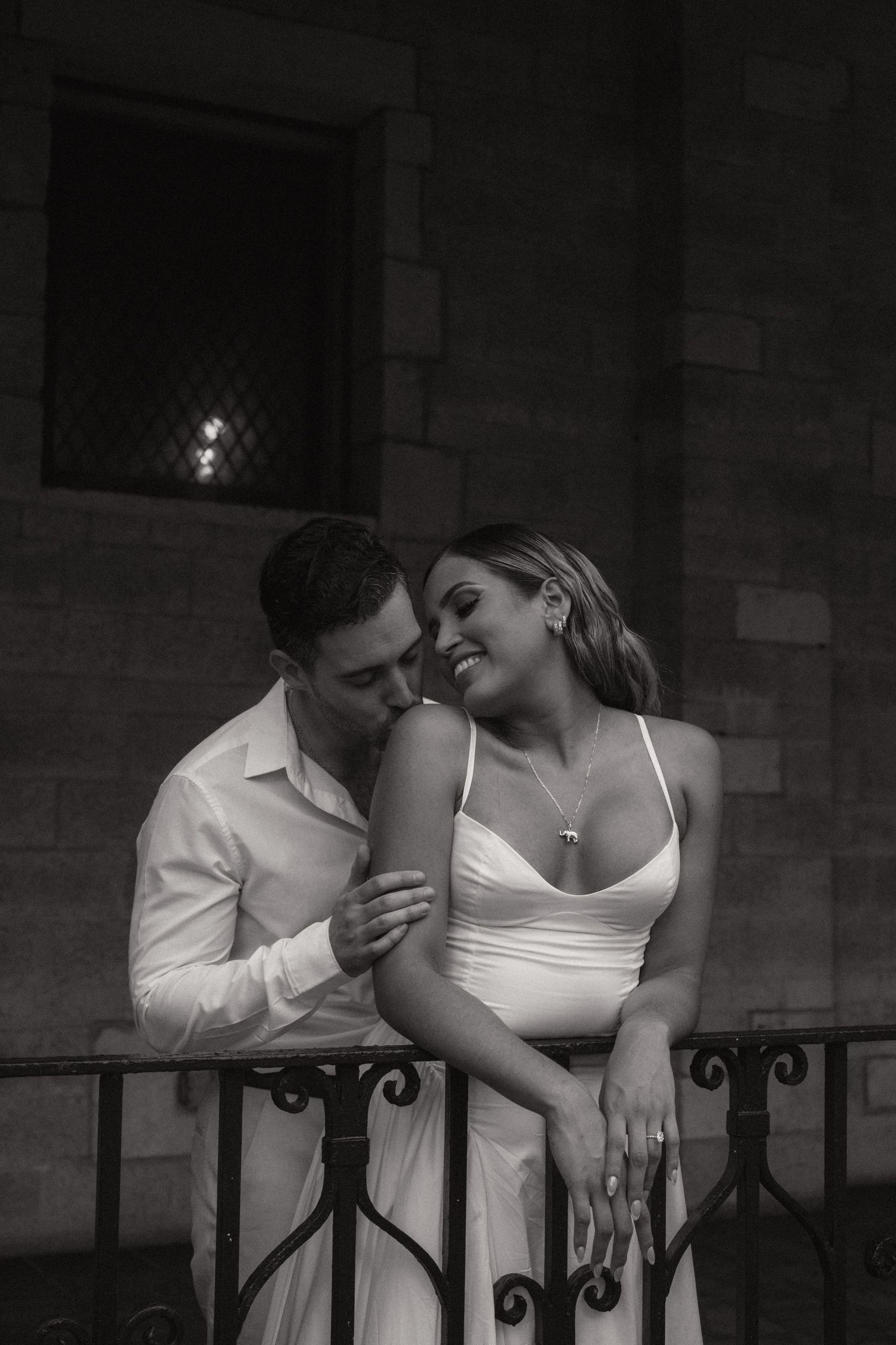 Black and white photo of a smiling woman in a white dress leaning on an iron railing as a man in a white shirt kisses her shoulder.