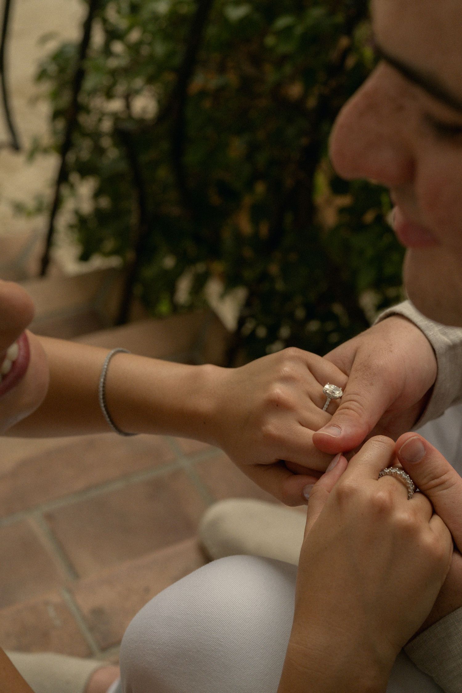 Close-up of two people holding hands, one wearing a diamond engagement ring and bracelet, with green plants in background.