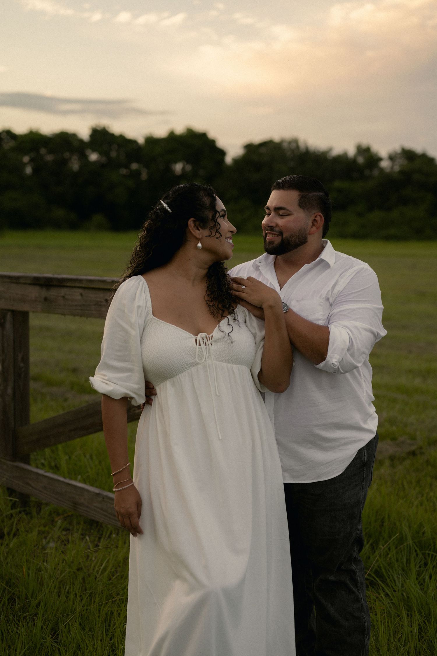 Couple in white outfits sharing a romantic moment in a green field at sunset near a wooden fence.